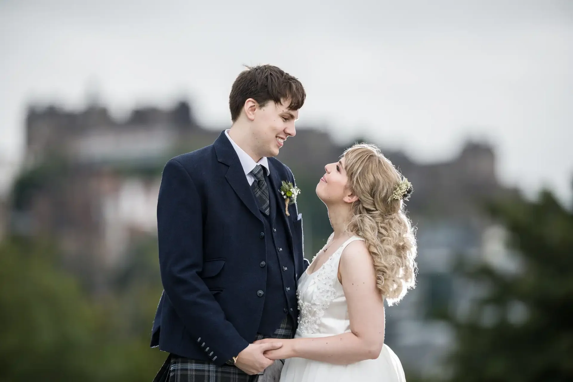 A bride and groom smiling at each other, wearing traditional scottish attire, with a castle in the background.