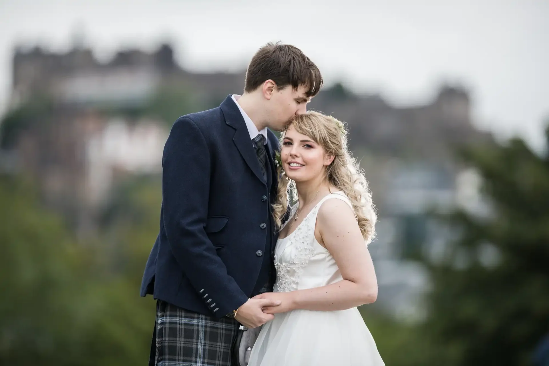 A newlywed couple in wedding attire embrace, with the groom kissing the bride's forehead, against a backdrop featuring edinburgh castle.
