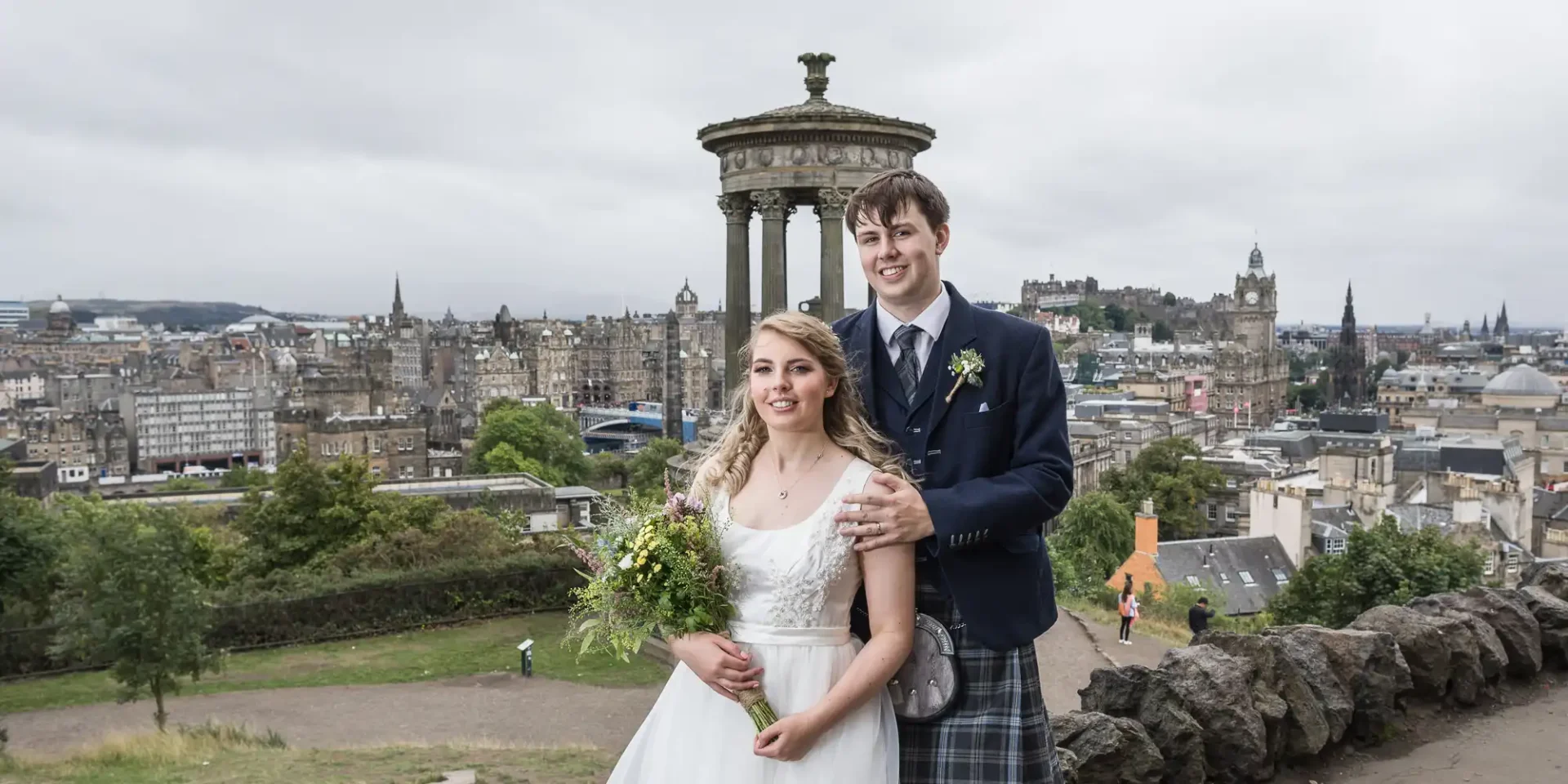 A newlywed couple posing with calton hill and edinburgh cityscape in the background. the groom wears a kilt, and the bride holds a bouquet.