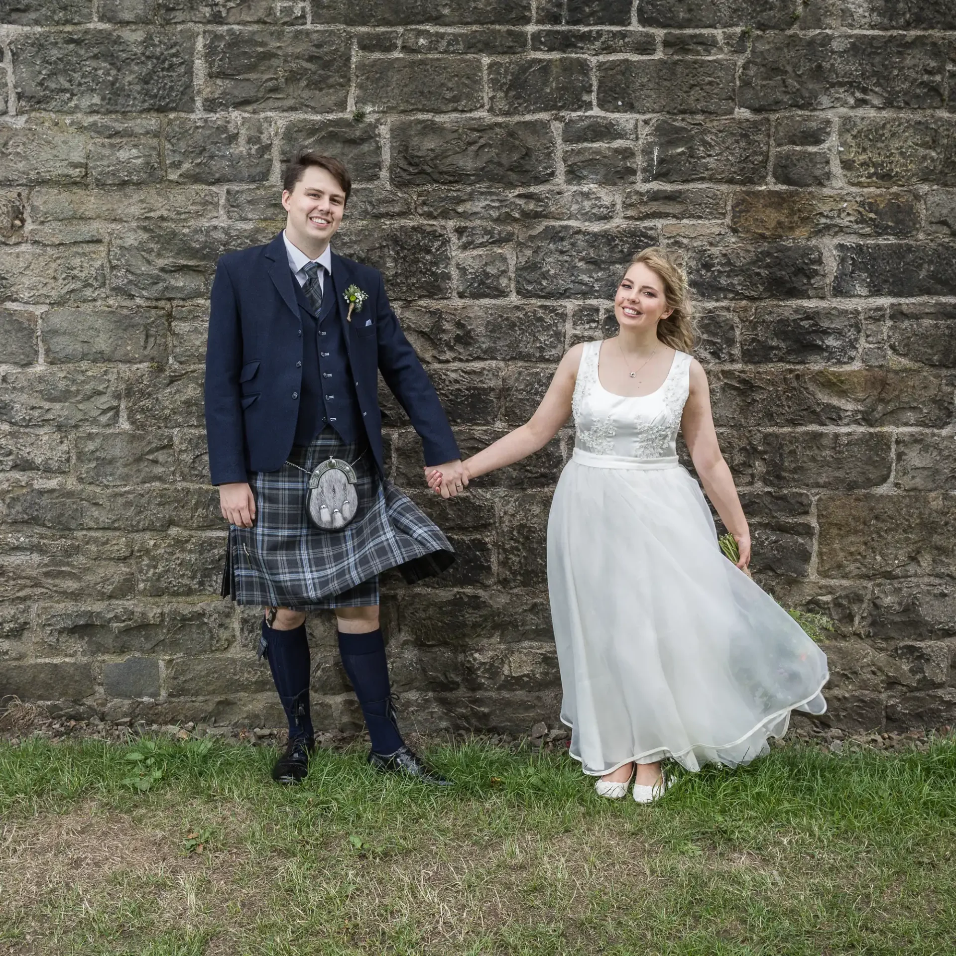 A bride and groom smiling and holding hands in front of a stone wall, the groom wearing a kilt and the bride in a white dress.