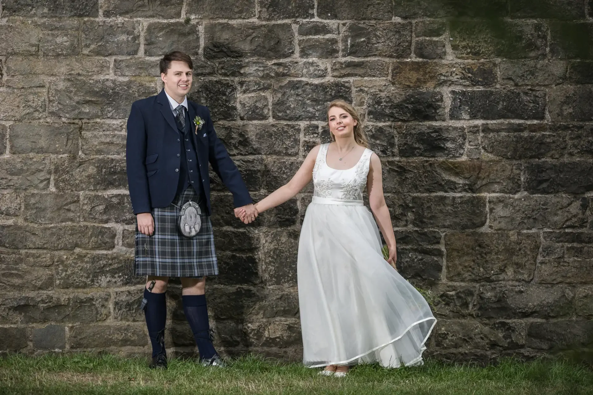 A bride in a white dress and a groom in a kilt holding hands in front of a stone wall, smiling contentedly.