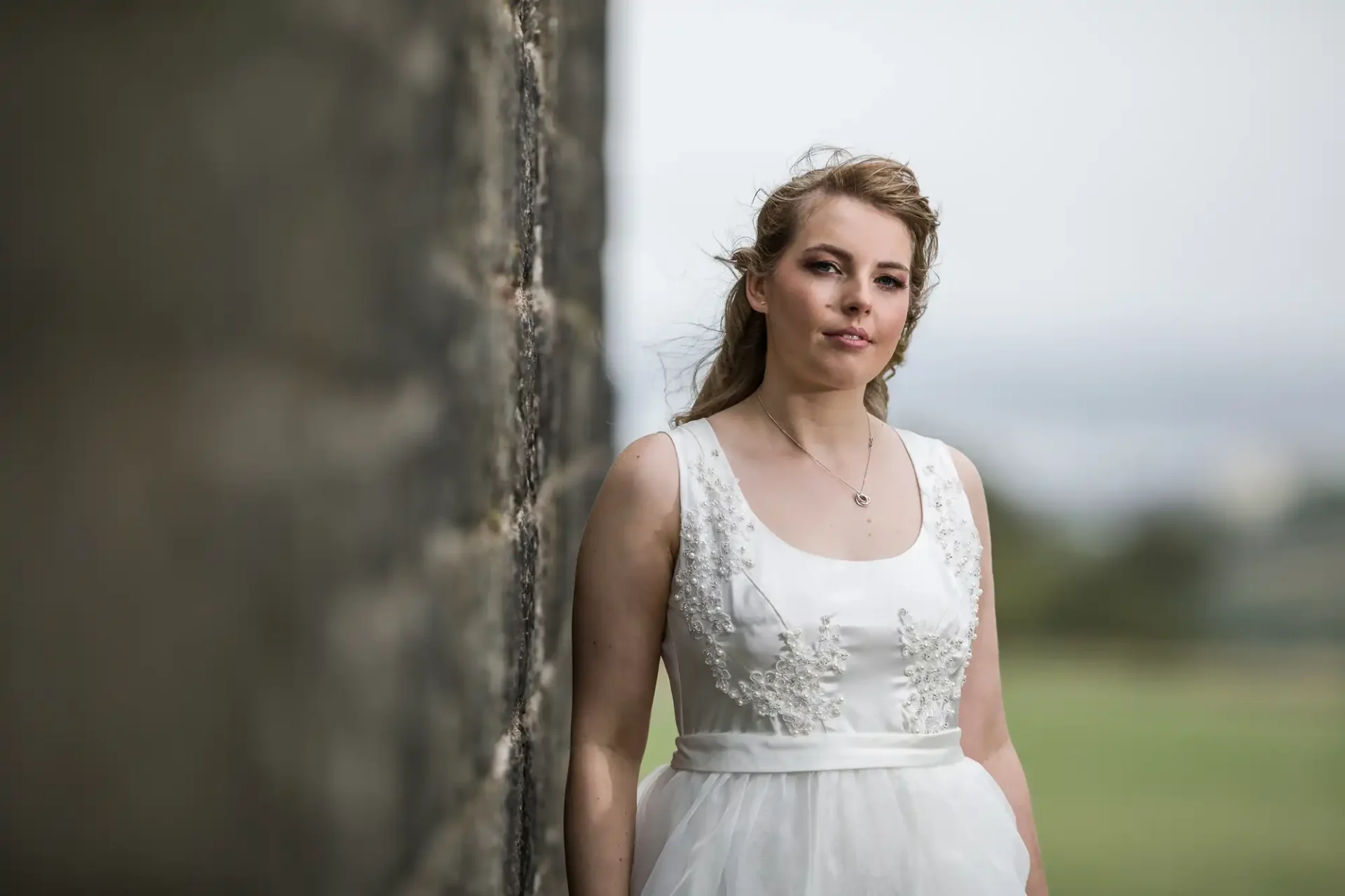 A woman in a white bridal gown stands beside a stone wall, her hair styled in an updo, looking pensively into the distance with a blurred natural background.
