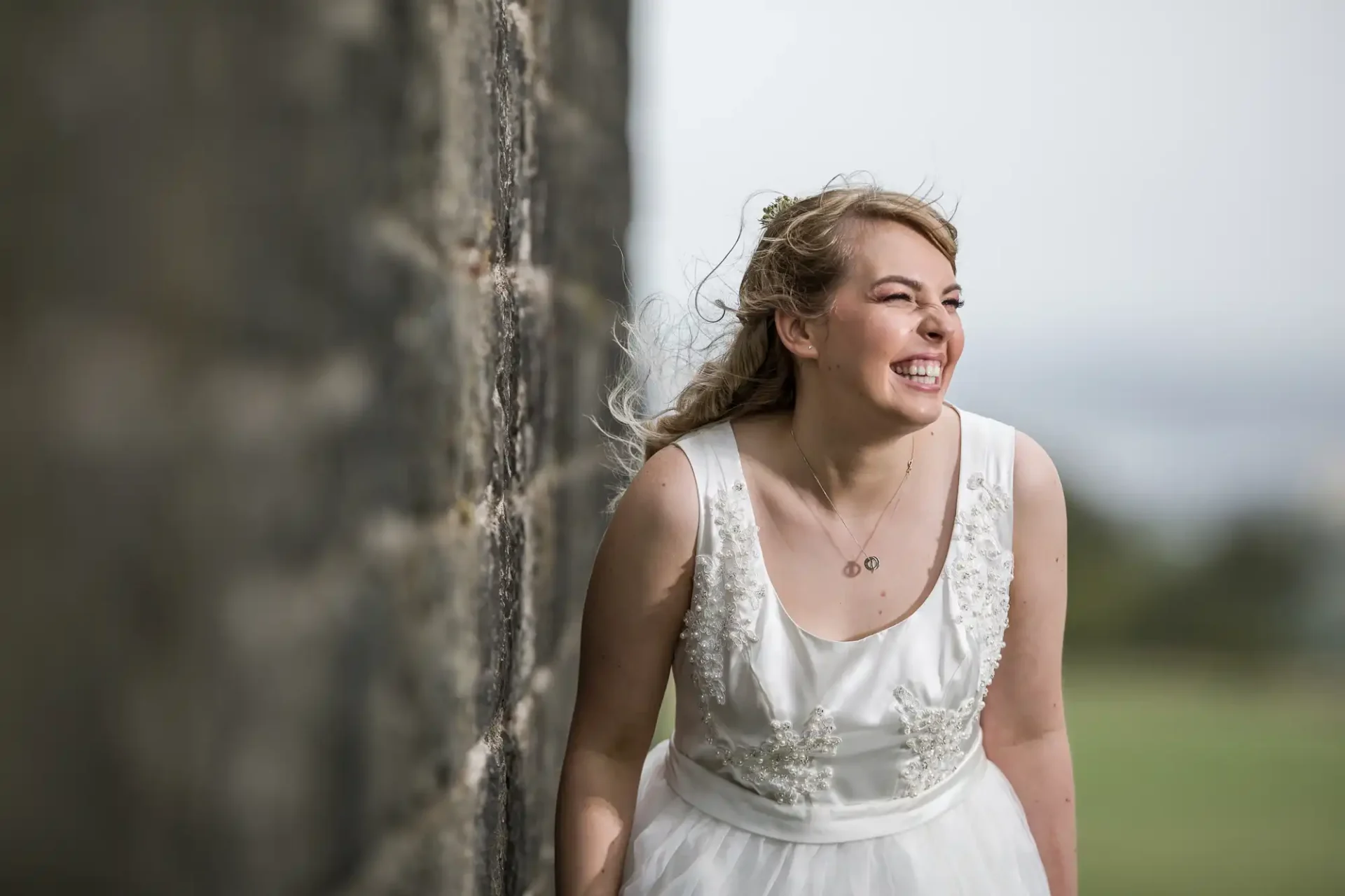 A joyful bride in a white dress with embellished details, smiling as she leans on a stone wall in a grassy outdoor setting.