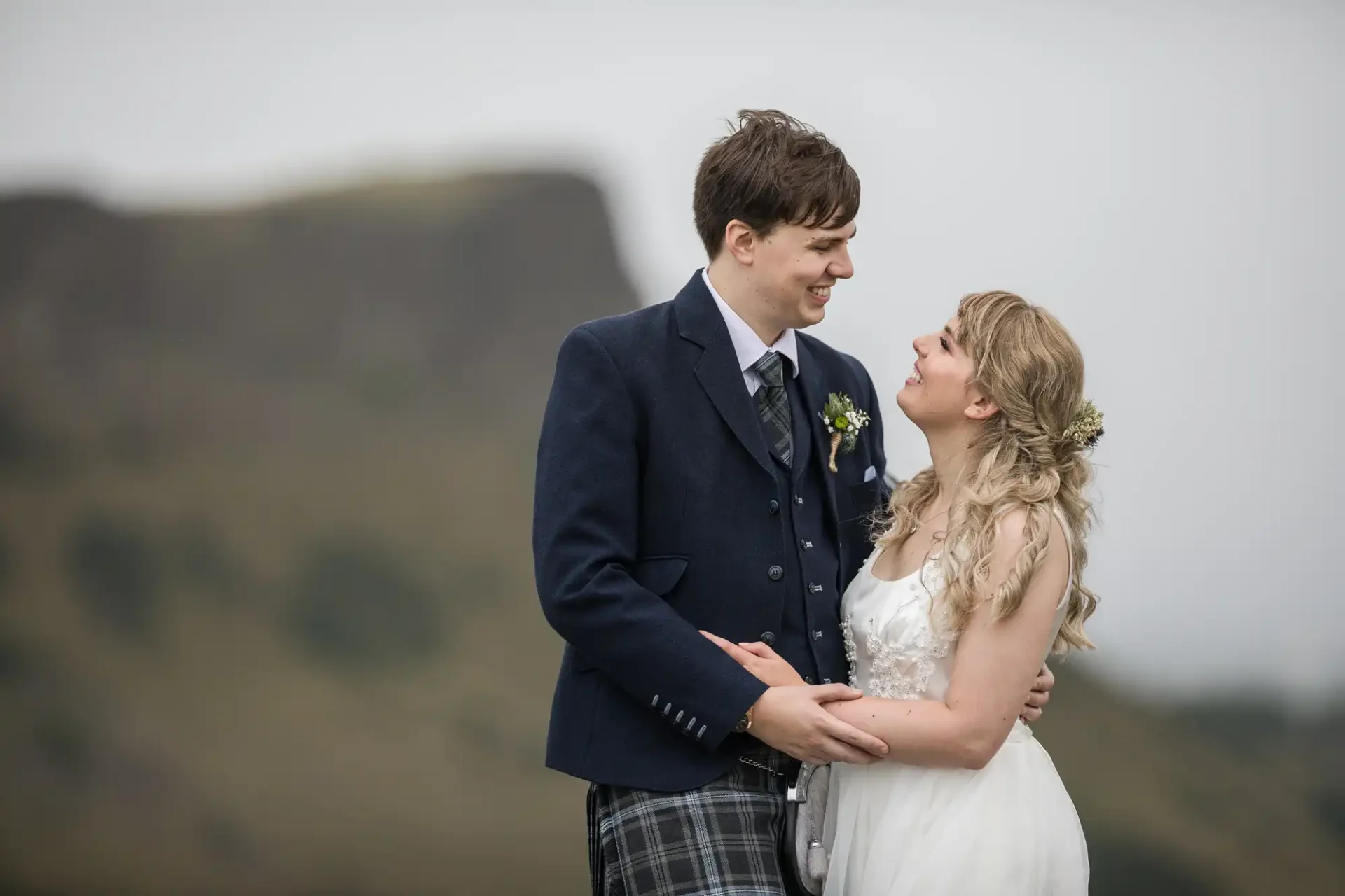 A bride and groom in wedding attire smiling at each other, standing outdoors with a blurred hill in the background.