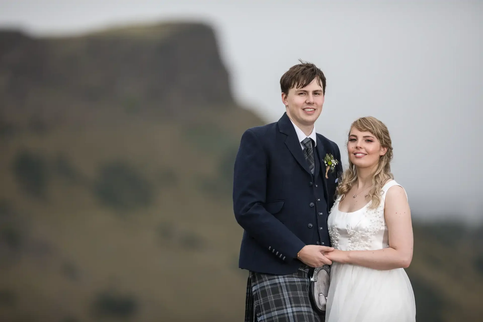 A bride and groom in formal attire smiling outdoors with a misty hill in the background.