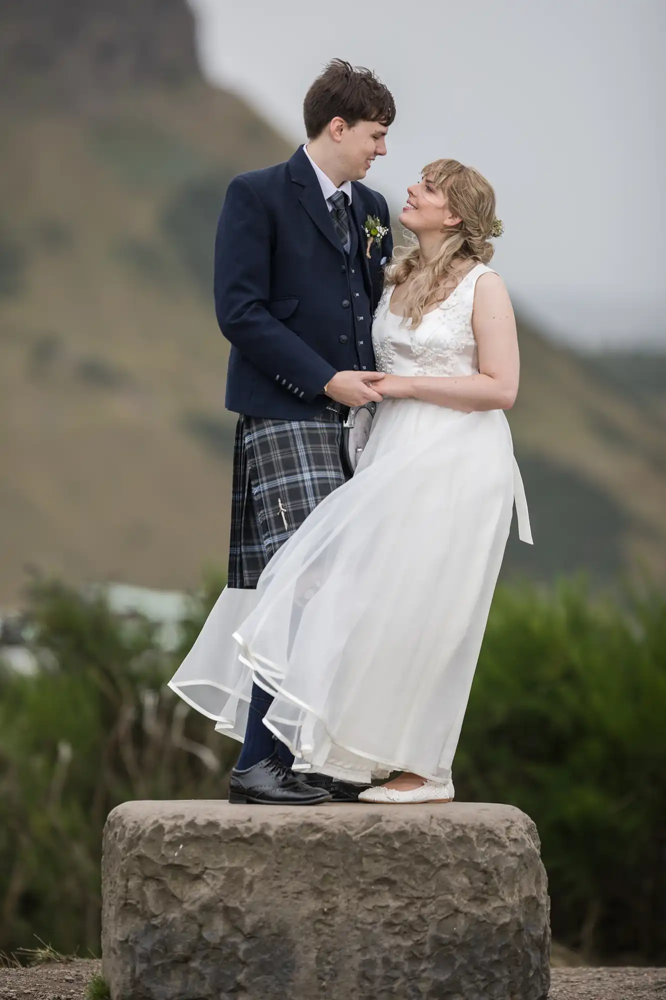 A bride and groom in wedding attire stand on a stone, facing each other with a mountainous backdrop.