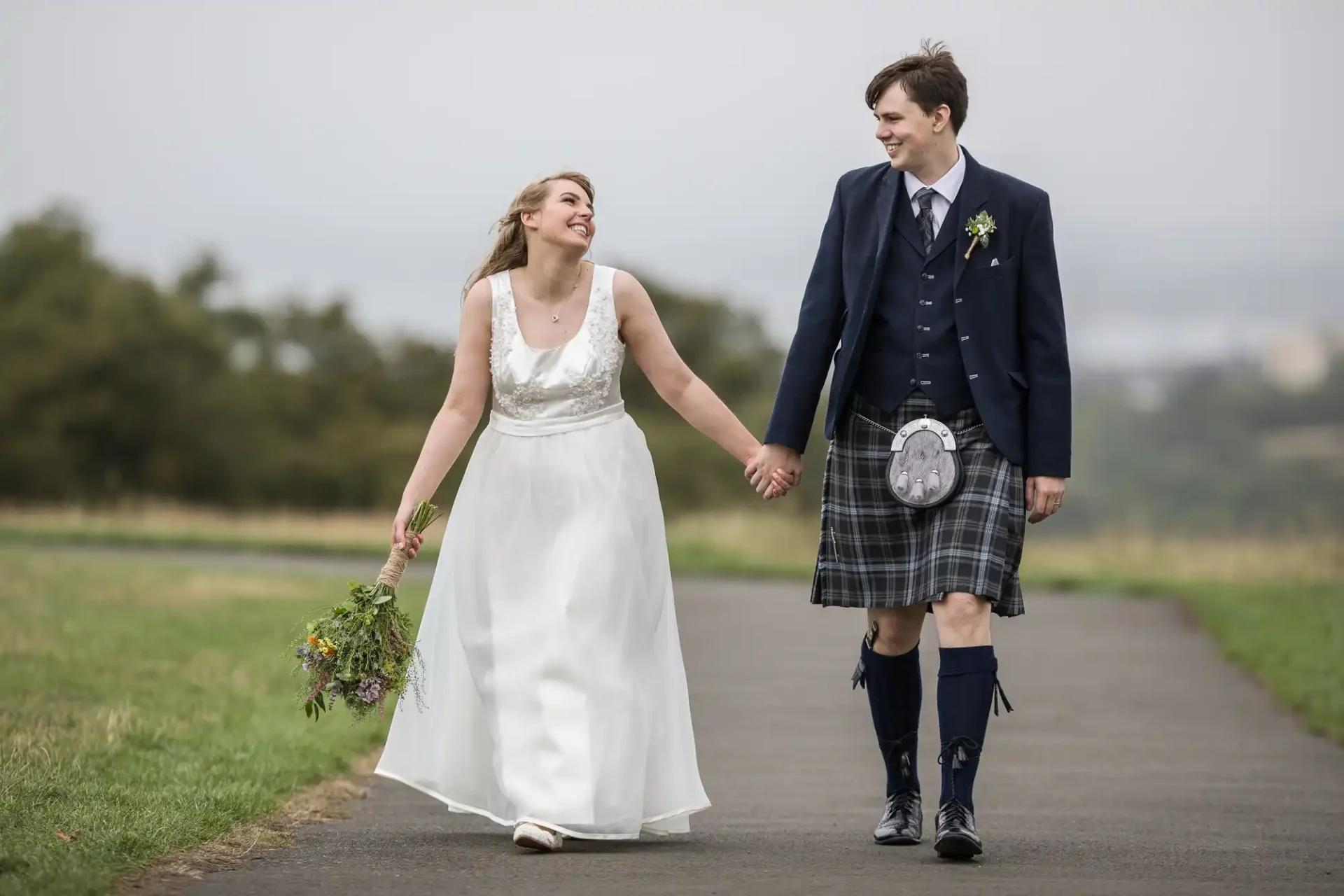 A newlywed couple walks hand in hand on a path, the bride in a white dress and the groom in a kilt, both smiling joyfully.