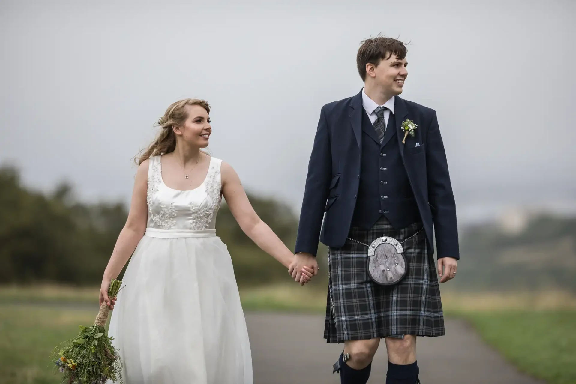 A bride and groom holding hands and walking on a pathway; the groom wears a kilt and the bride carries a bouquet, with a grassy field in the background.