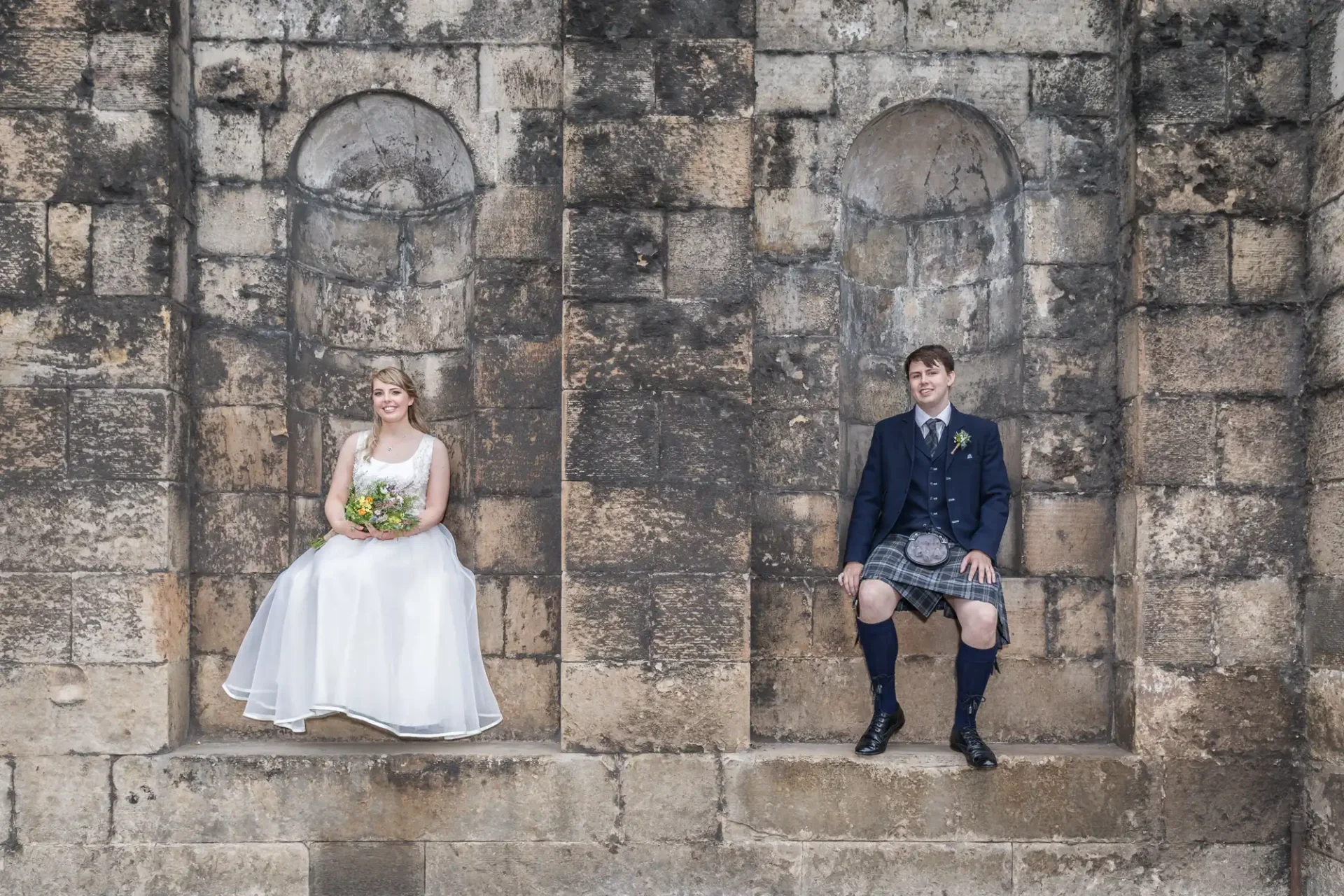 Bride and groom in formal wear sitting separately in two stone niches of an old wall, smiling.
