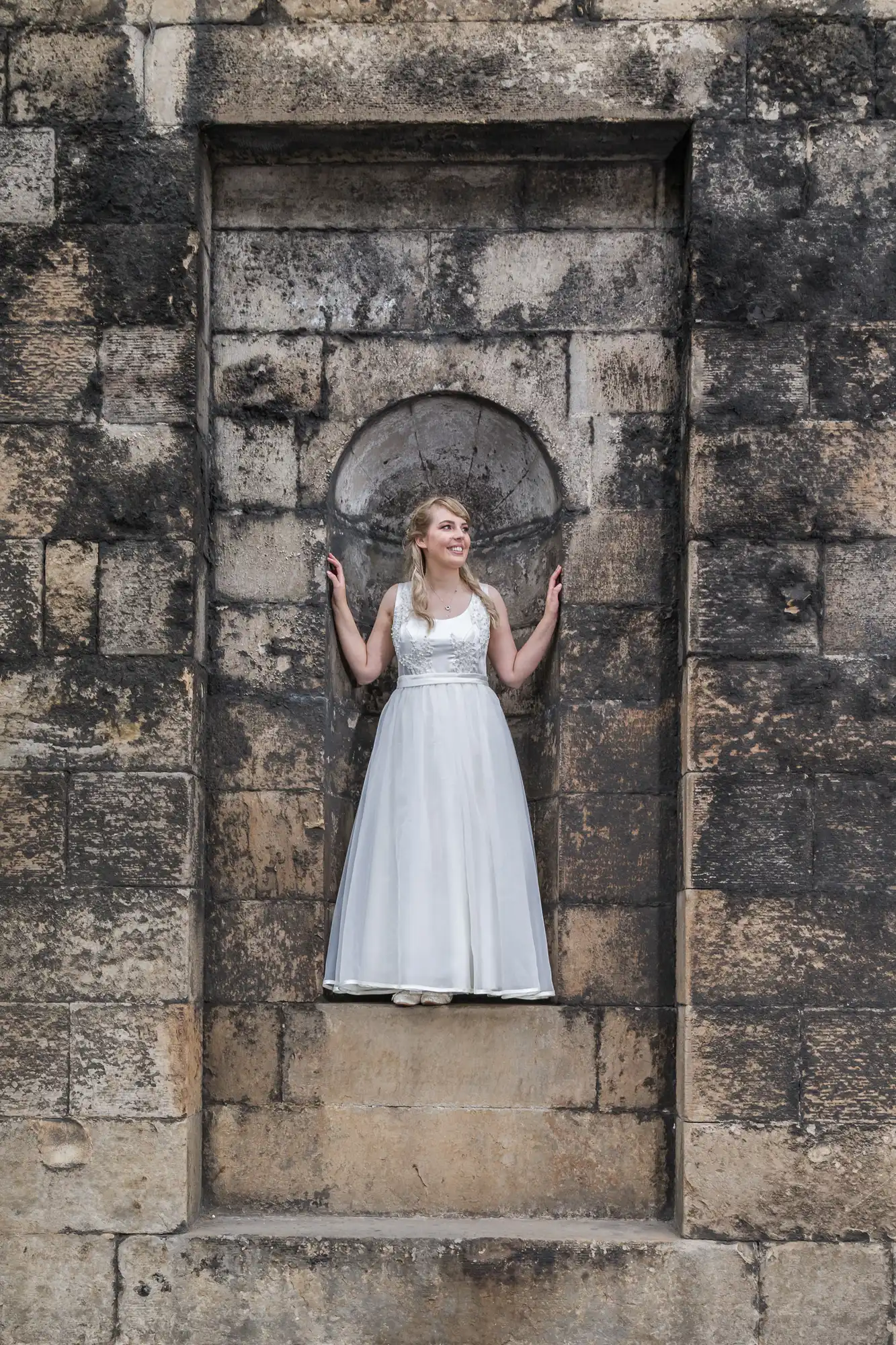 A woman in a white dress stands smiling in a large stone niche with her arms raised slightly, in an old textured wall.