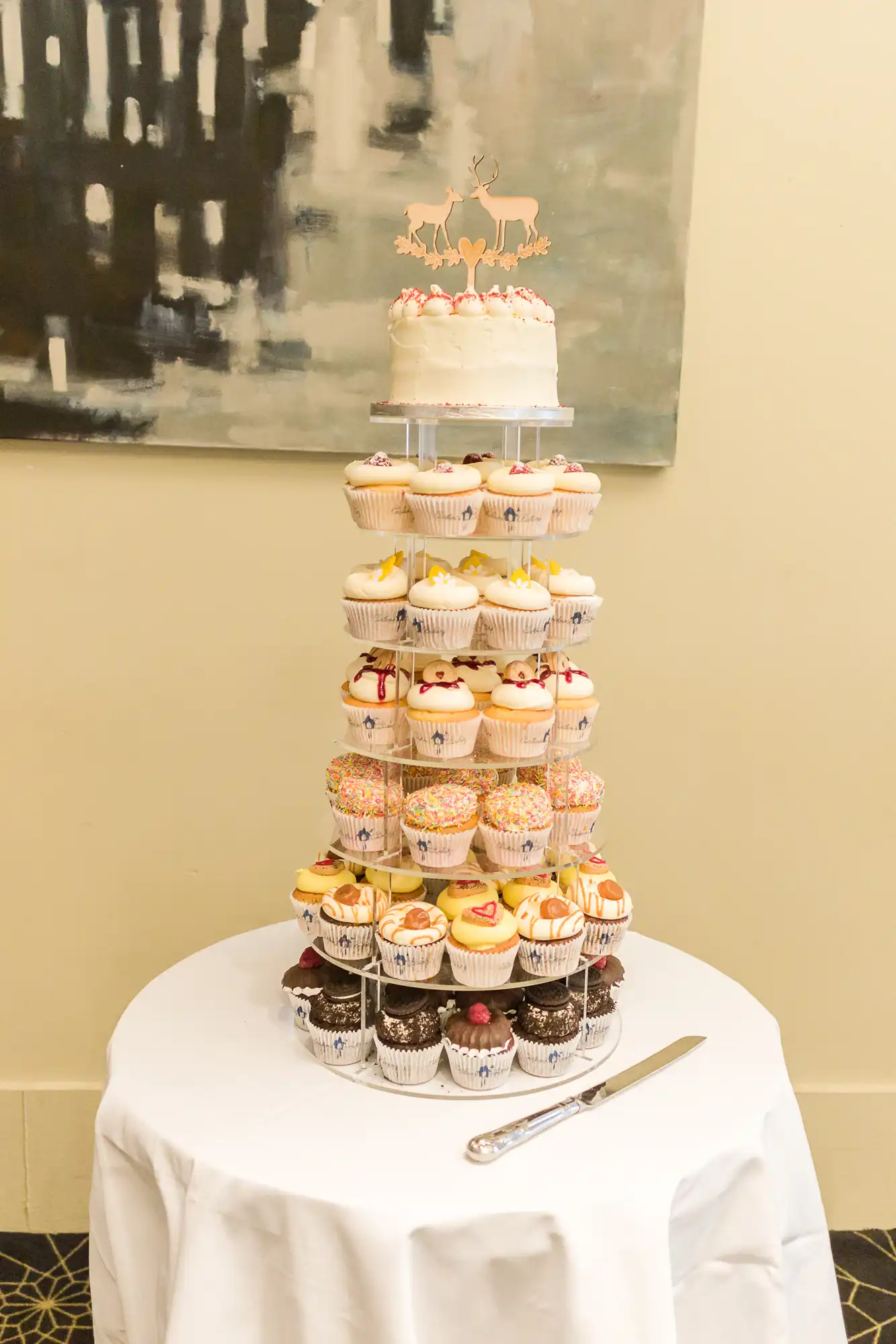 A tiered cupcake stand with various frosted cupcakes topped by a small wedding cake, displayed under an abstract painting in a warmly lit venue.