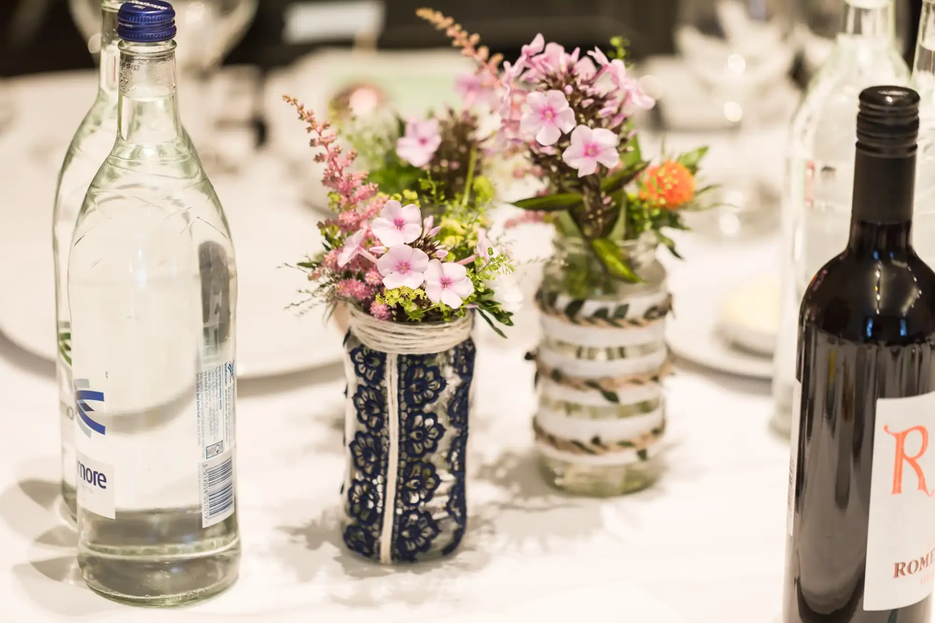 A table setting featuring a clear water bottle, a wine bottle, and small bouquets in decorative vases.