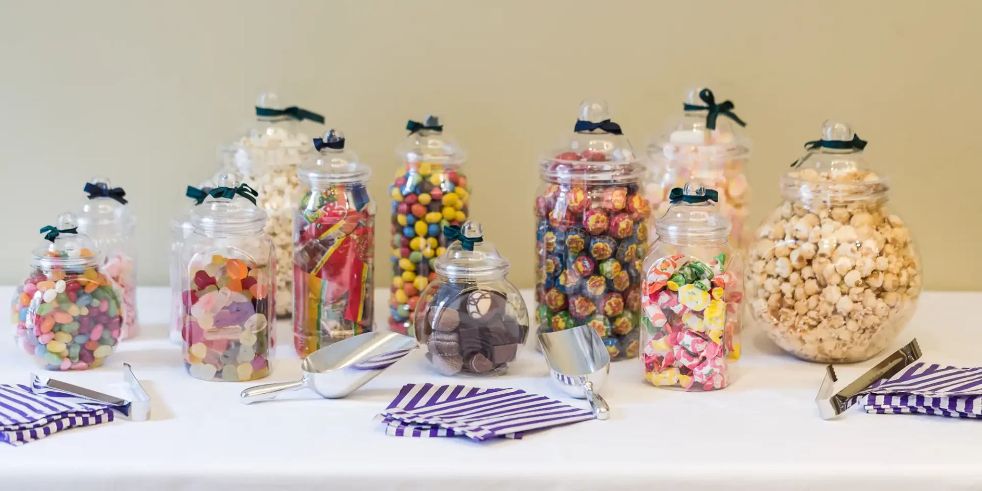 A variety of candies in glass jars arranged on a table with metal scoops and striped napkins.