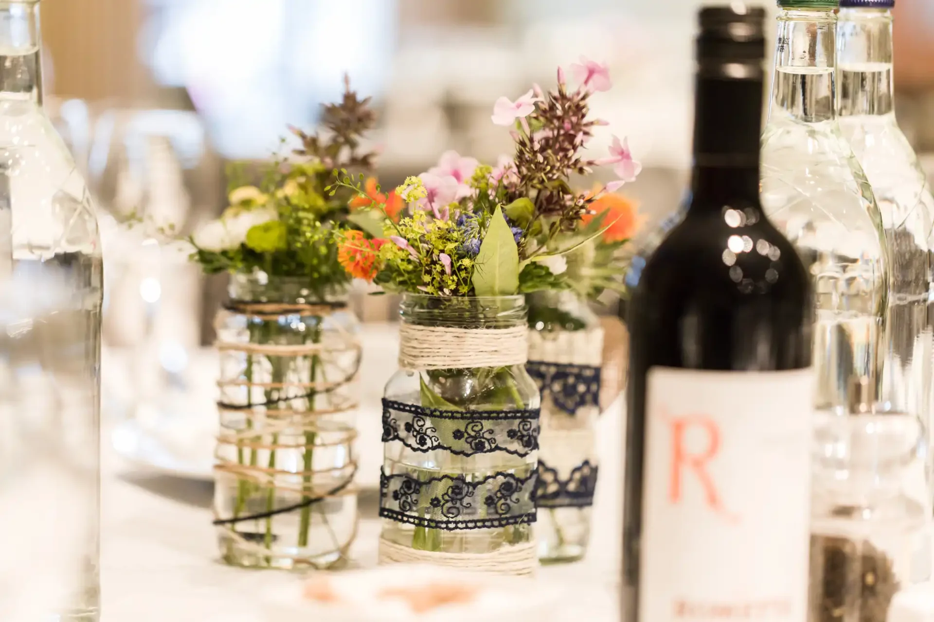 Table setting with small floral arrangements in glass jars, a water bottle, and a wine bottle labeled with the letter 'r'.
