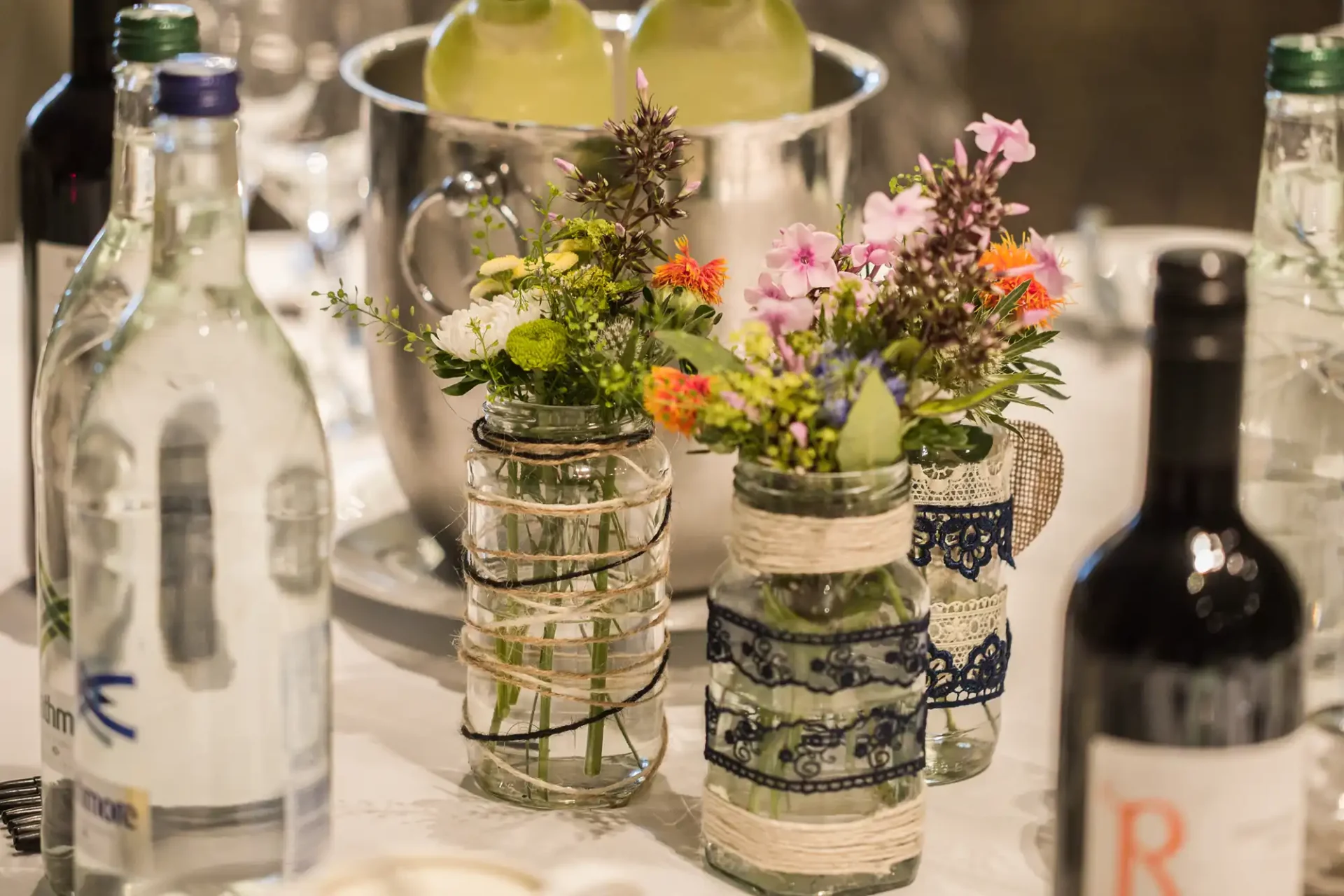 Table setting featuring small floral arrangements in glass jars, surrounded by bottles of water and wine, with a backdrop of dishware.