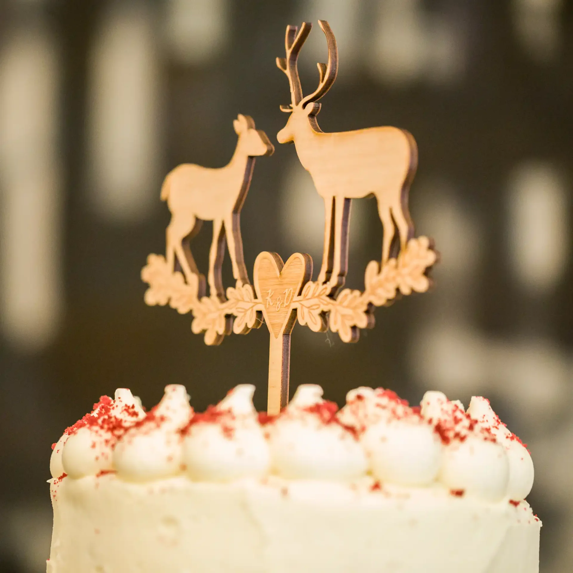 A close-up of a cake topper featuring two deer figures surrounded by a heart, on top of a cake with white icing and decorative red sprinkles.