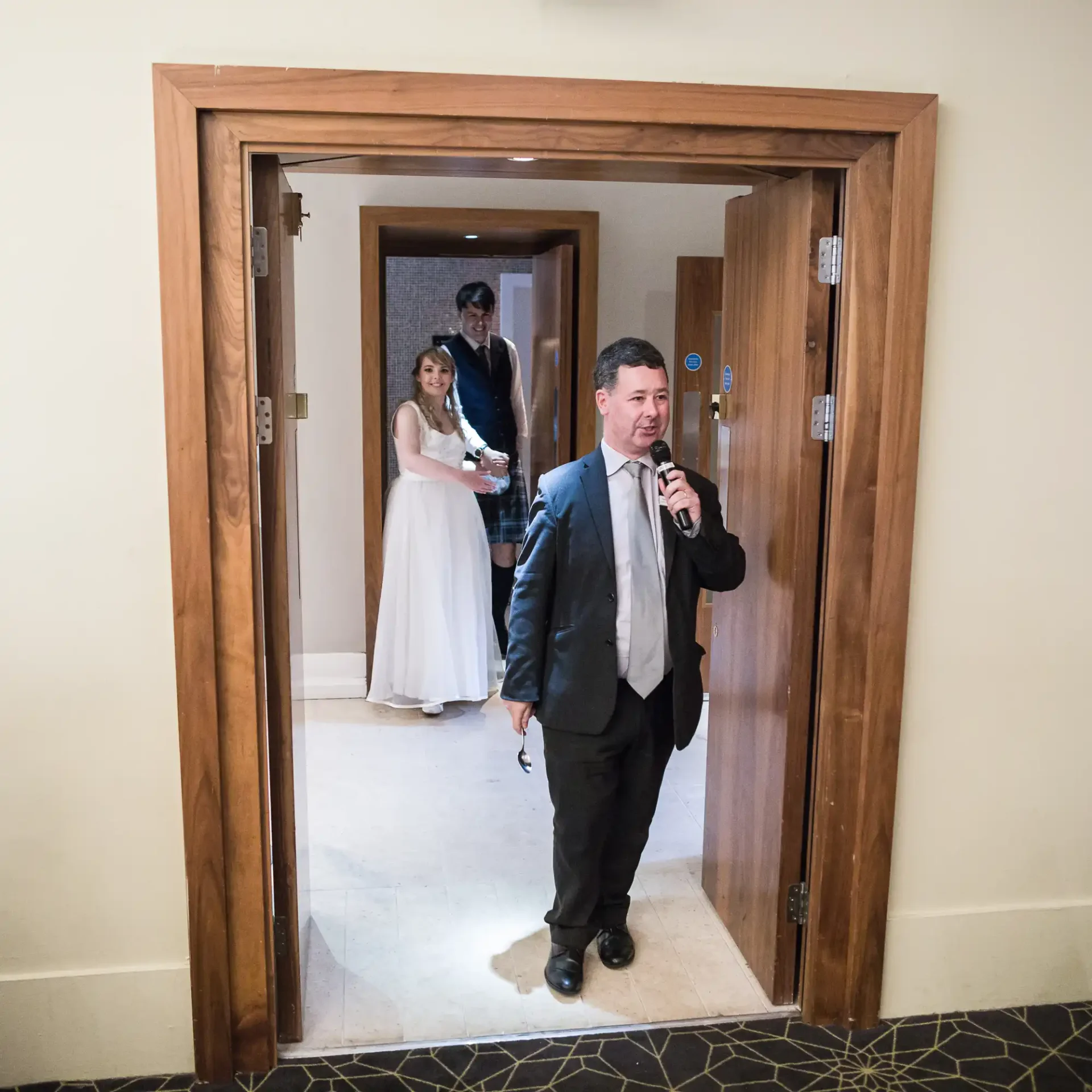 A man in a suit exits a room talking into a microphone, while a couple in wedding attire stands inside the doorway behind him.