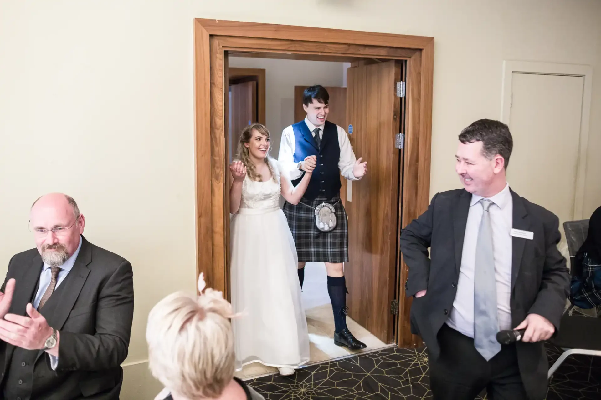 Bride and groom entering a room, the groom wearing a kilt, greeted with applause by guests.