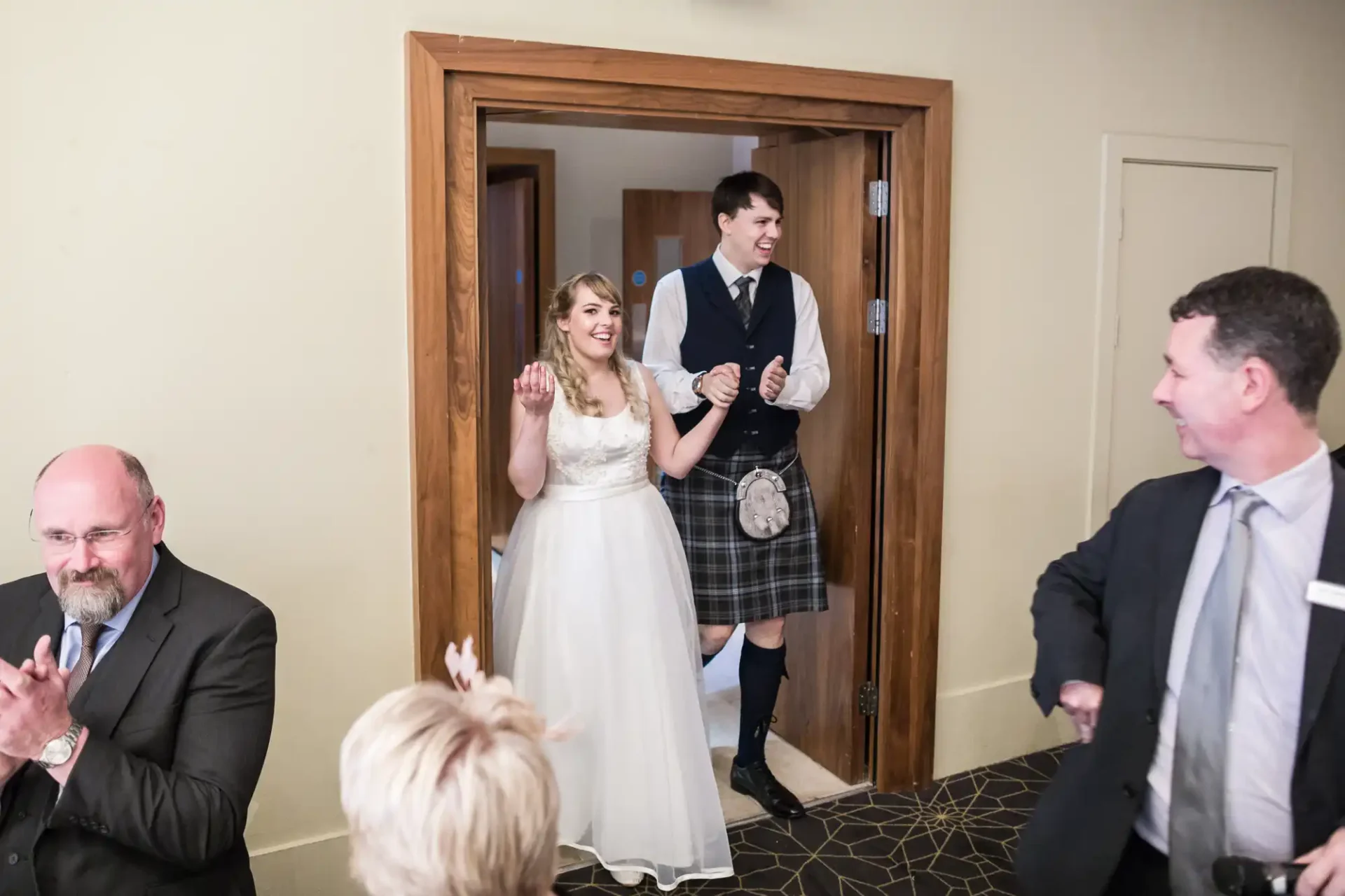 A bride and groom smiling as they enter a room, with guests clapping around them. the groom is wearing a kilt.