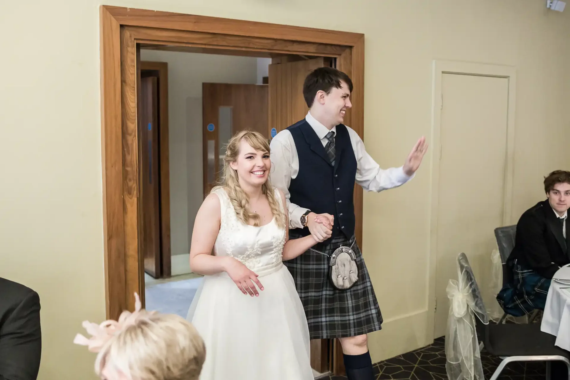 A bride and groom smiling and walking through a doorway, the groom in a kilt and the bride in a white dress.