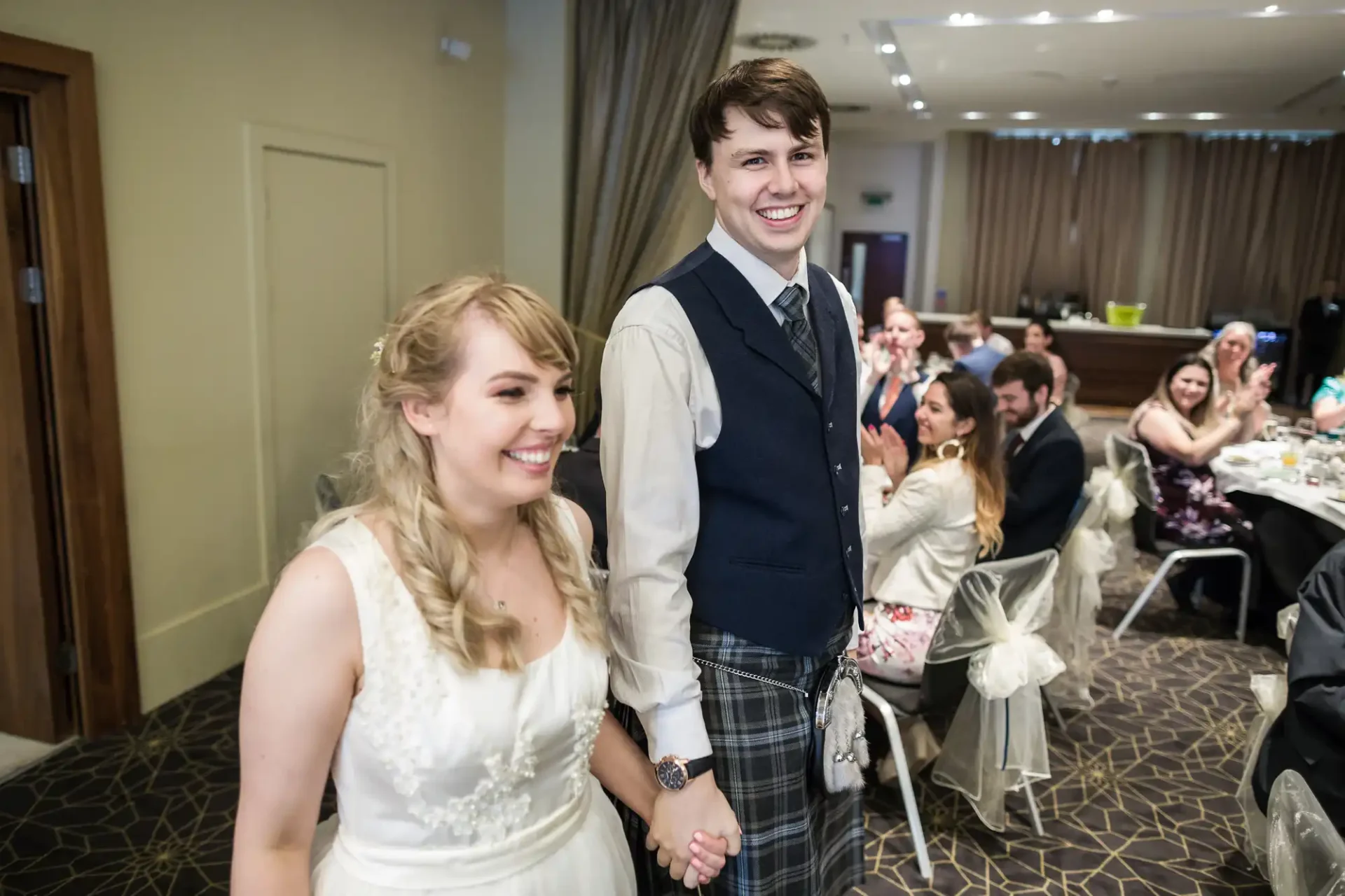 A bride and groom smiling as they walk hand in hand through a banquet hall with seated guests watching.