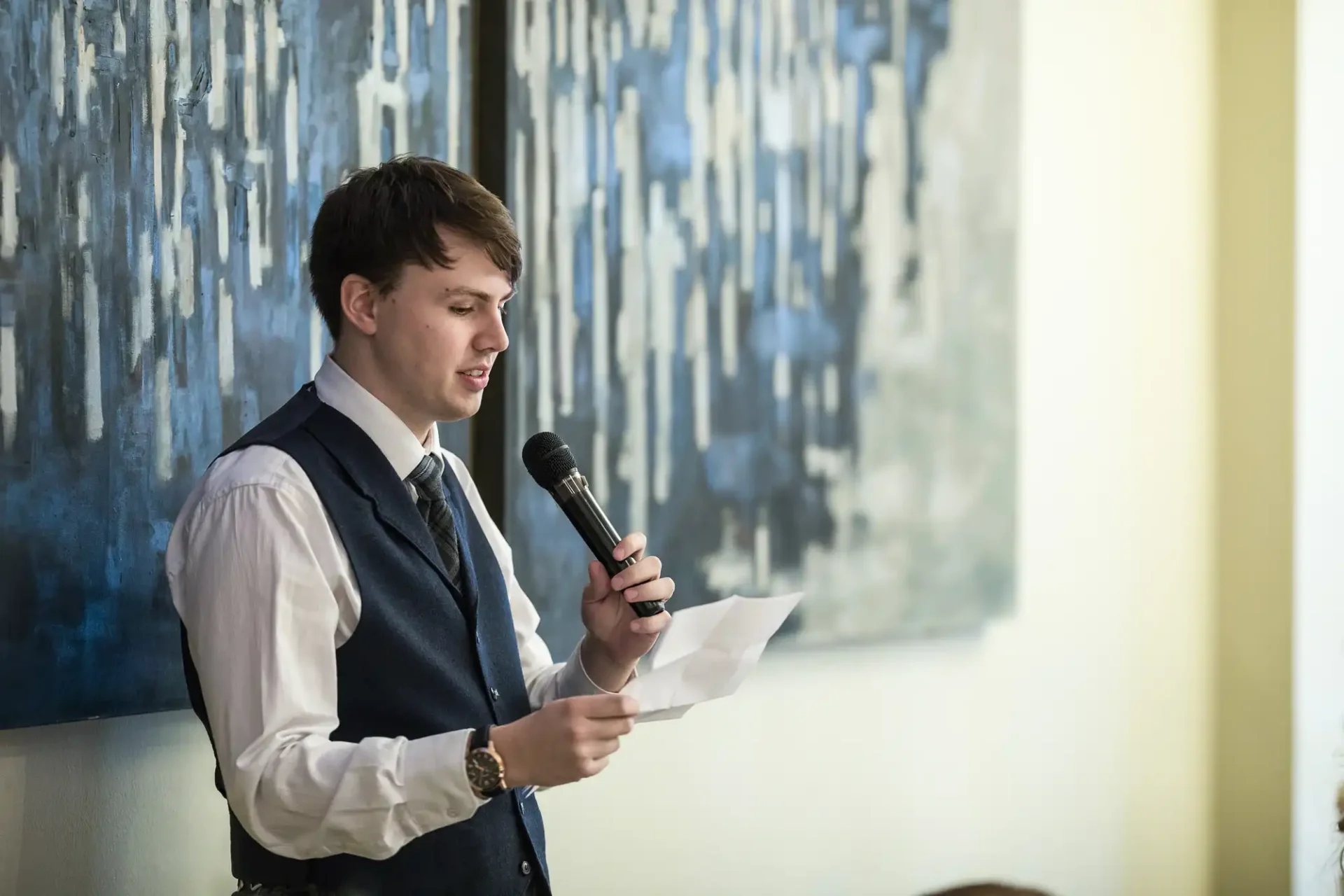 A young man in a vest and tie reading from a paper and speaking into a microphone against a backdrop of a blue abstract painting.