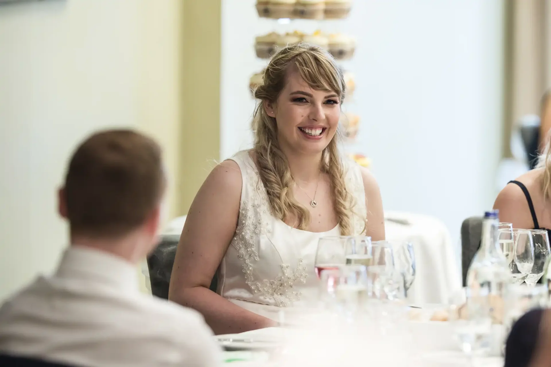 A bride smiling at a table during a wedding reception, surrounded by guests and table settings.
