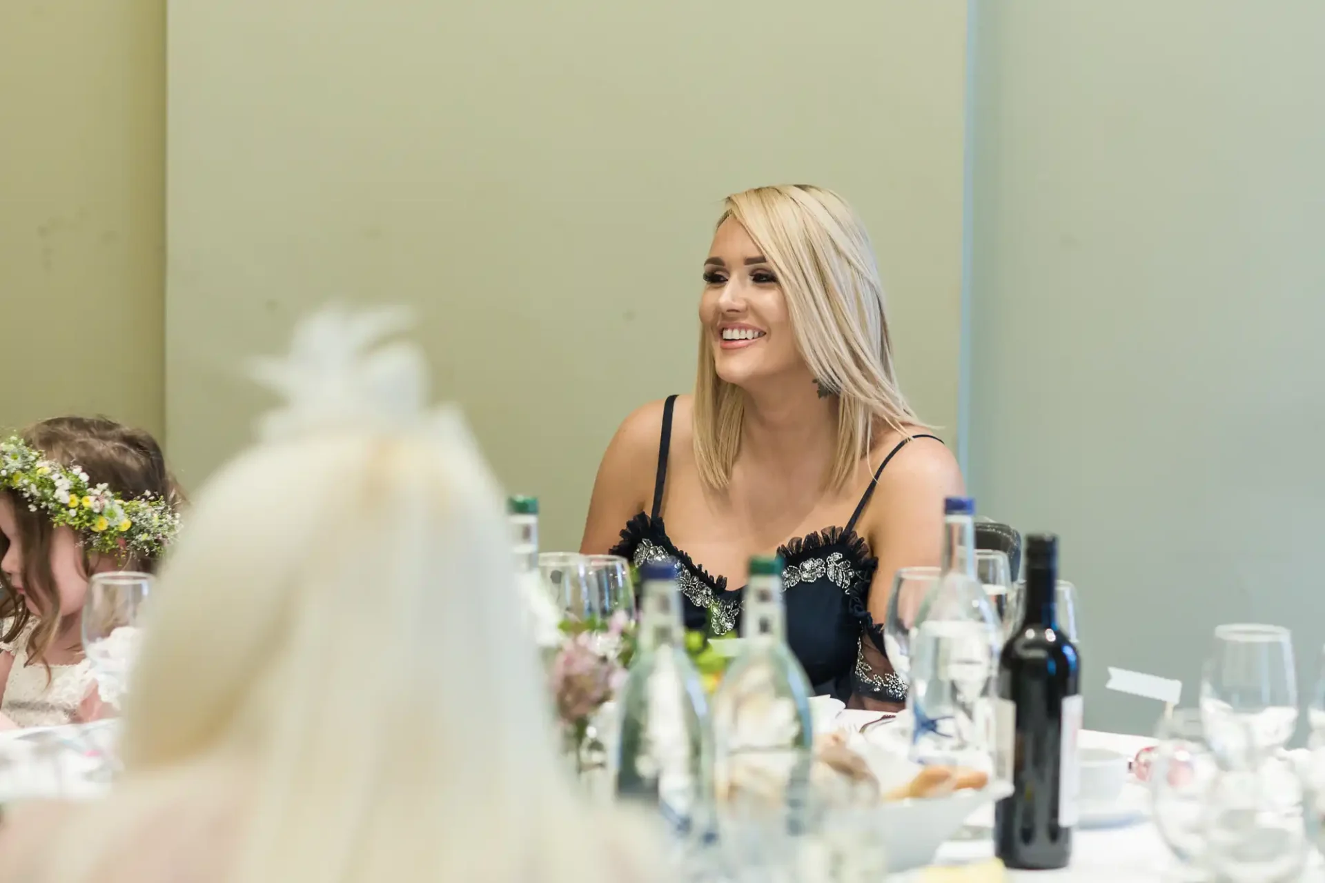 A woman with blond hair smiling at a table during a formal event, surrounded by wine glasses and guests.