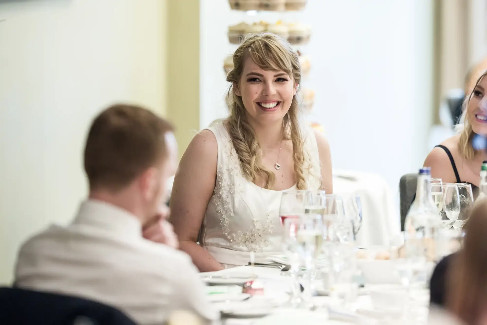 A bride smiling at a wedding reception table while guests interact around her.