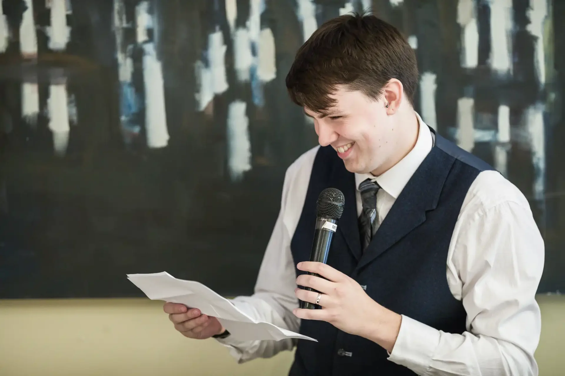 A young man in a vest and tie smiling while reading from a paper and holding a microphone.