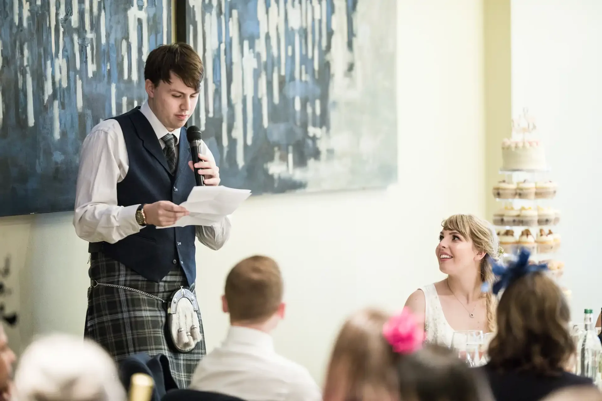 A man in a kilt and vest reads from a paper into a microphone at a wedding reception, with a smiling woman in a white dress listening nearby. a tiered cake stands in the background.