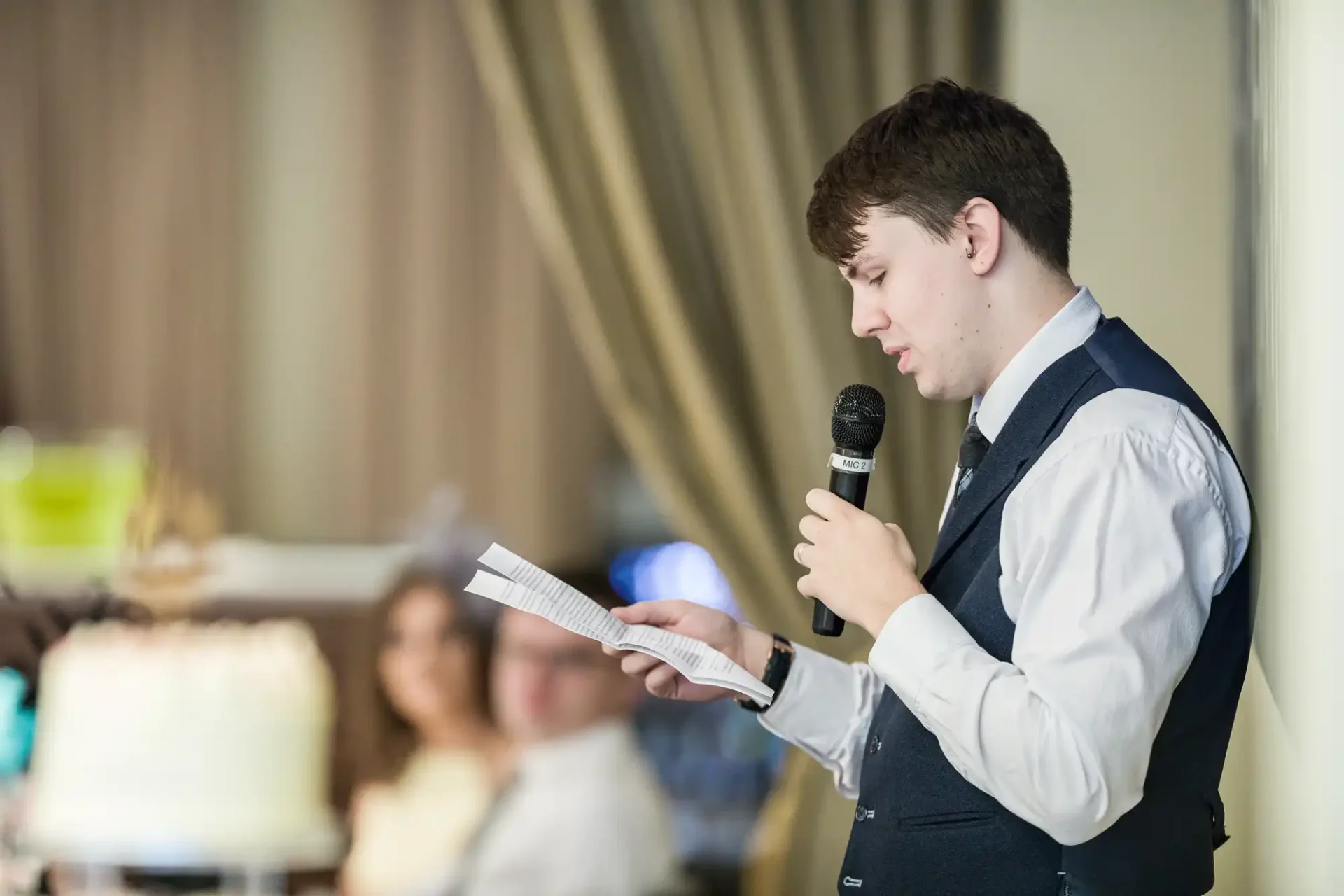 A young man in a vest and tie speaks into a microphone while reading from a paper at an indoor event.