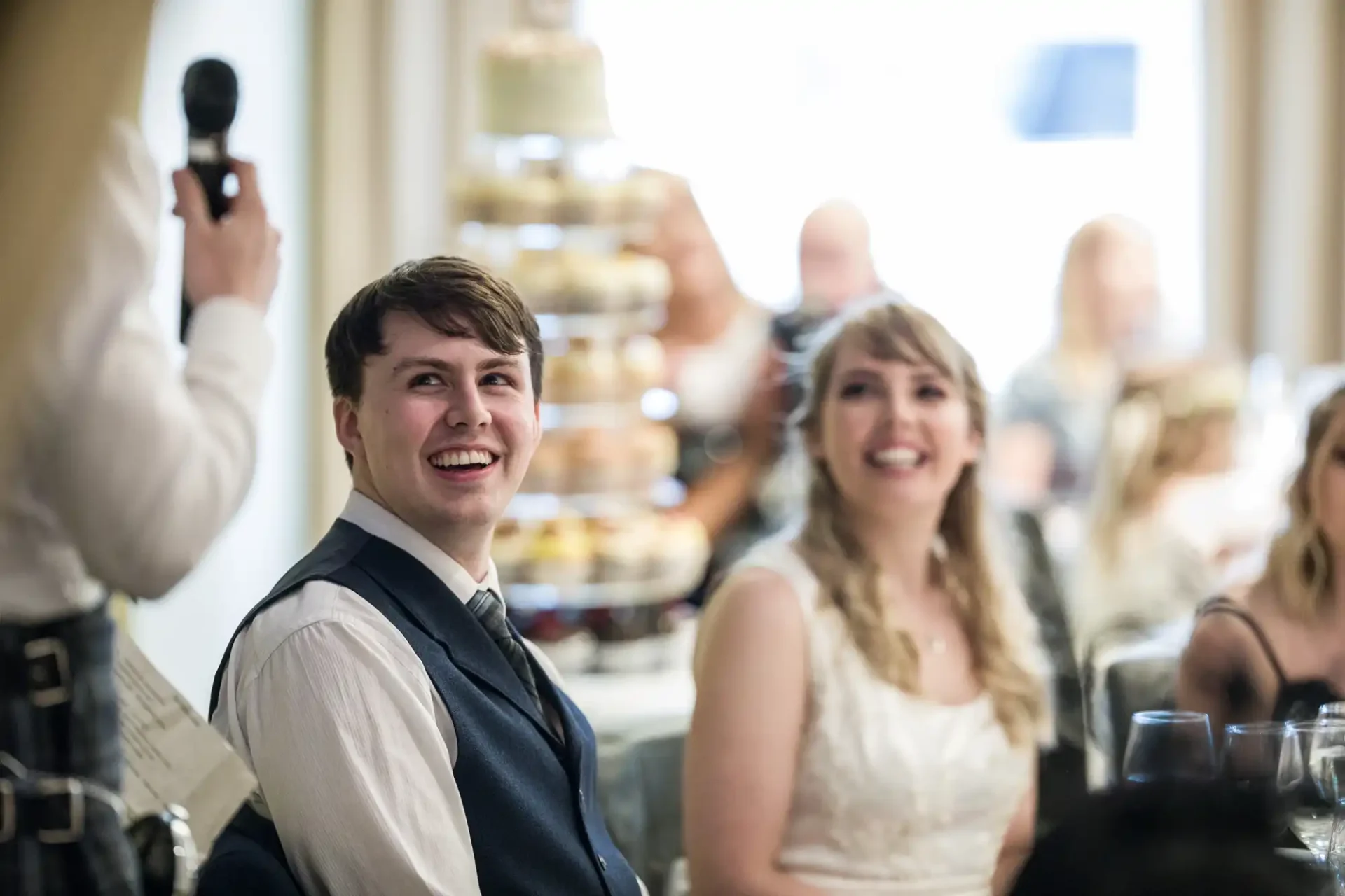 A joyful bride and groom seated at a wedding reception, smiling as a guest speaks with a microphone in the background.