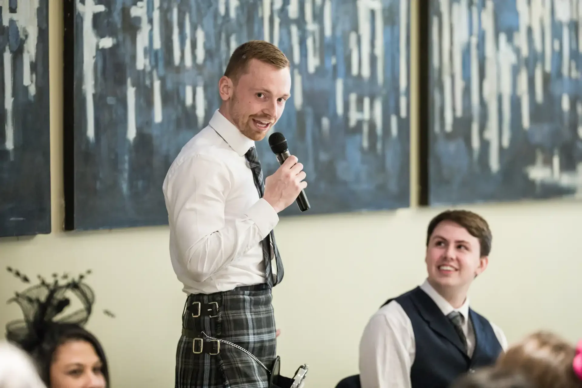 Man in a kilt and white shirt speaking into a microphone at an event, with another person smiling in the background.