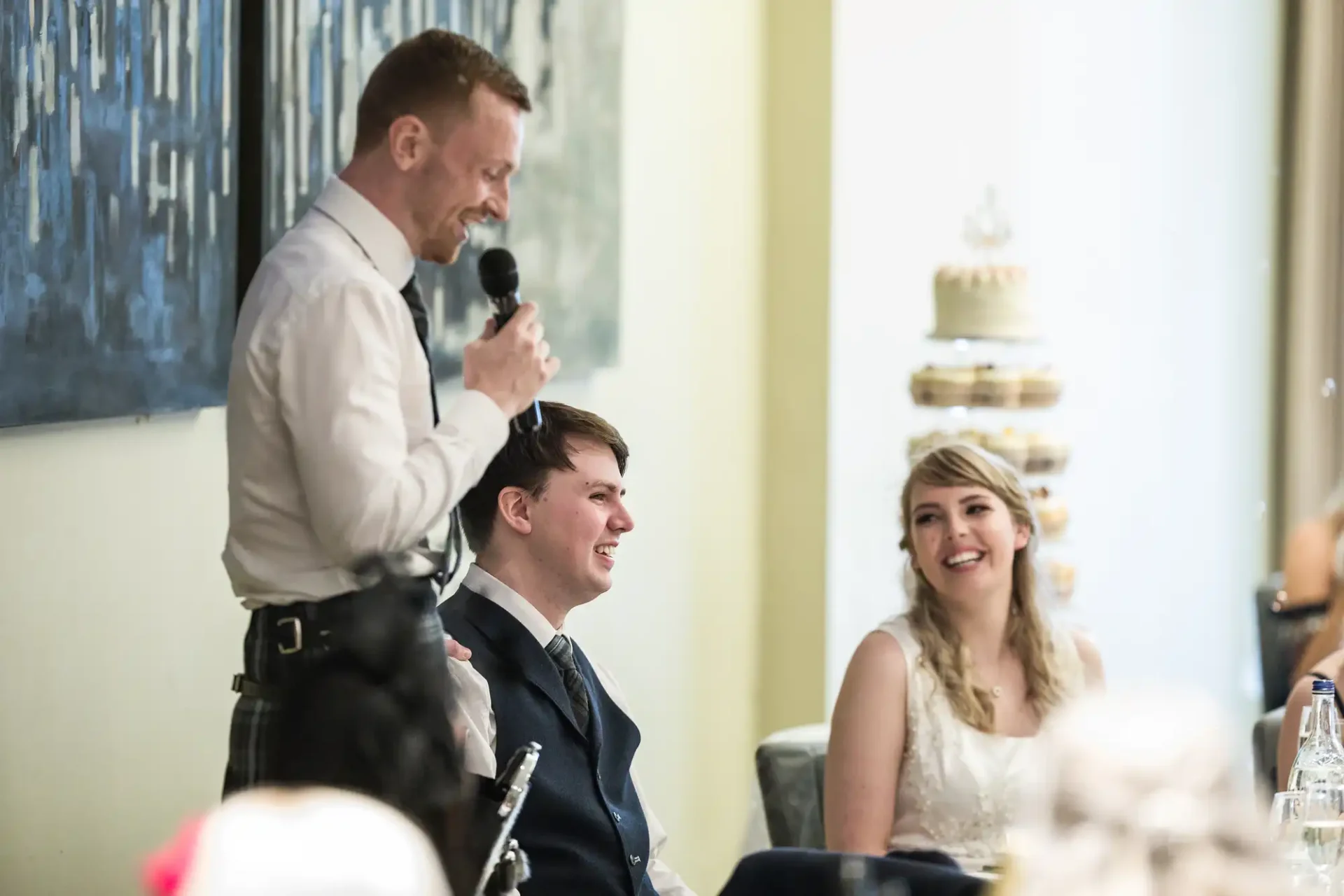 A man speaking into a microphone at a wedding reception, standing beside a seated bride and groom who are smiling.