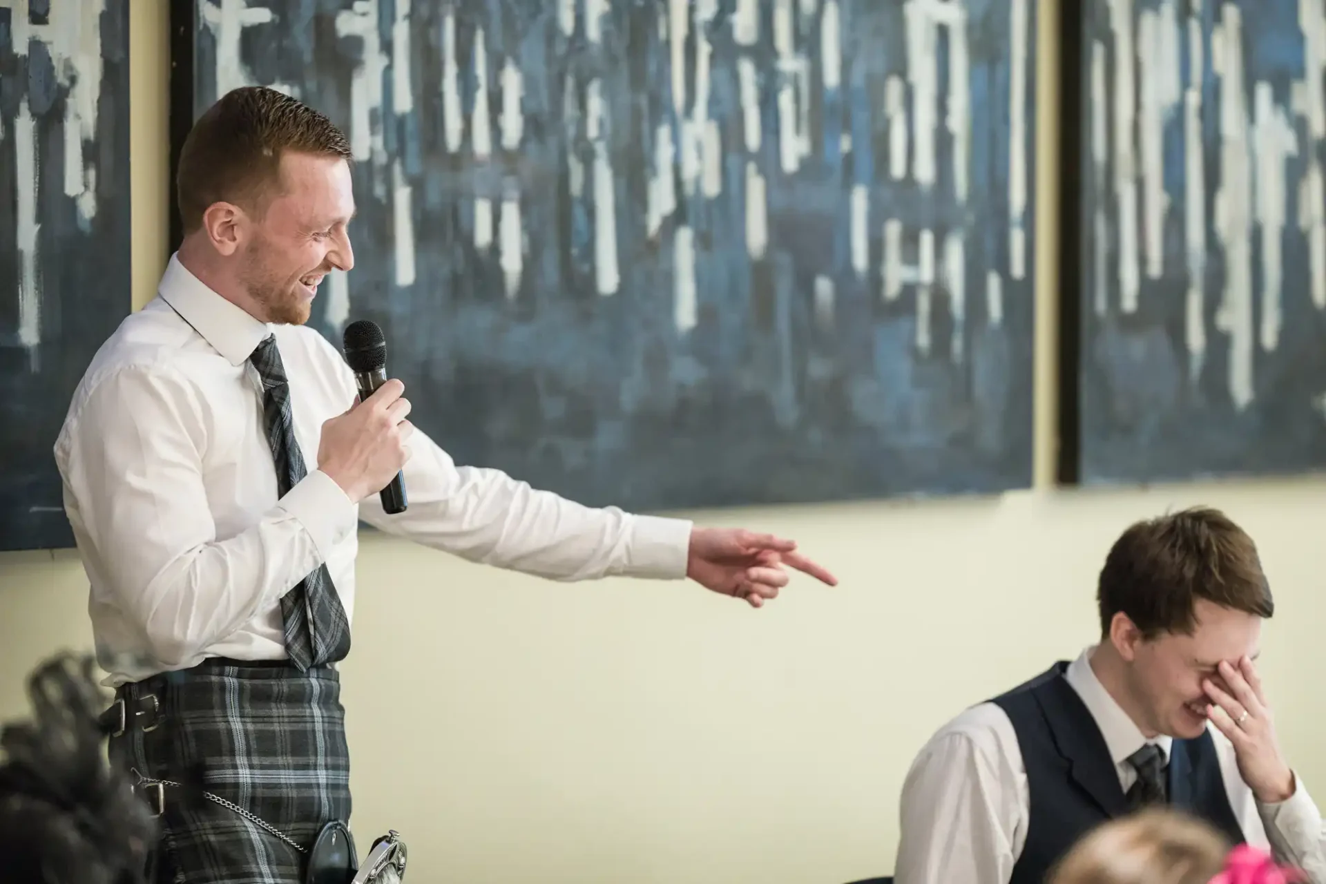 Man in plaid trousers and vest speaks into a microphone, gesturing towards another man who is laughing, in a room with abstract art on the wall.