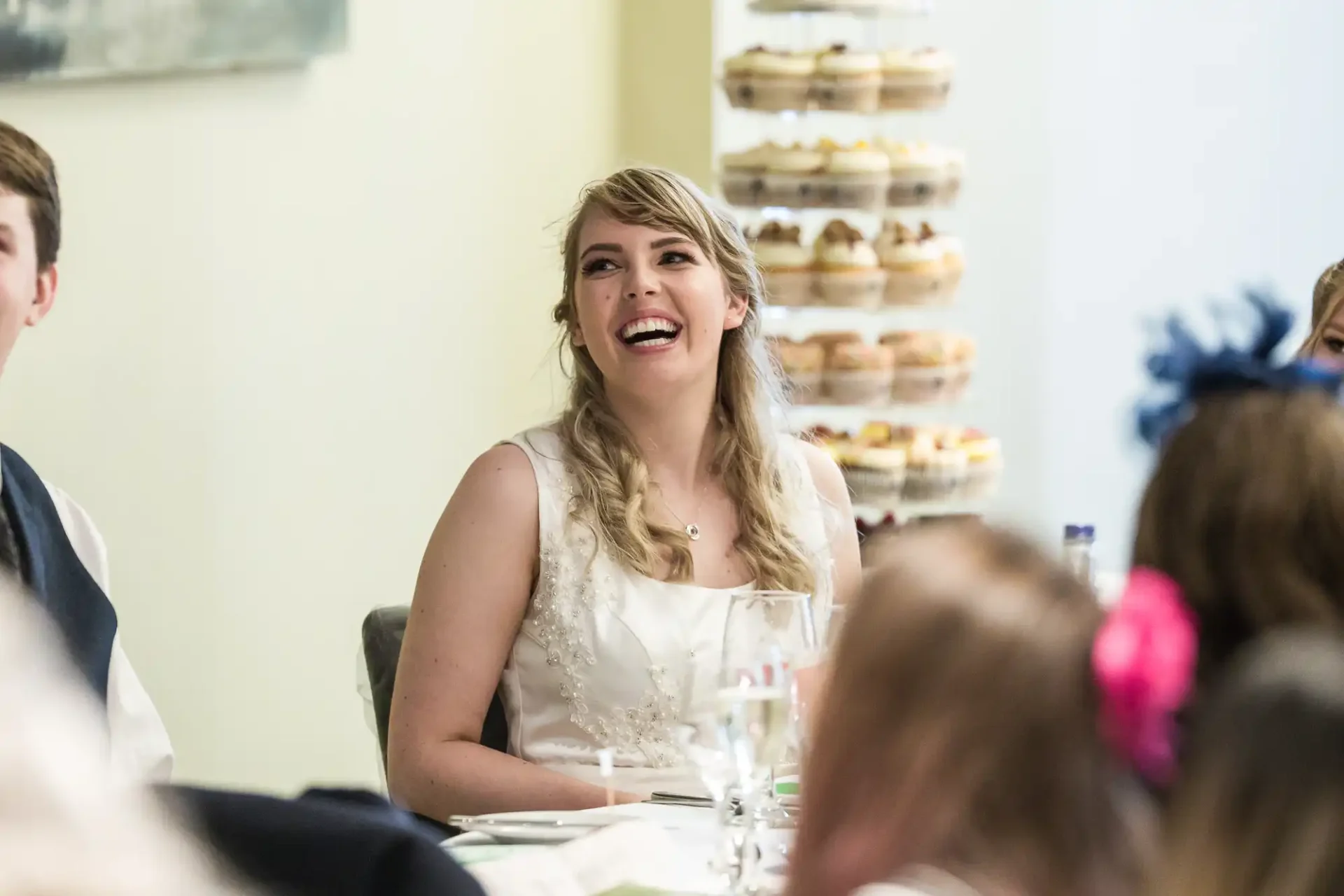 A bride smiling at a wedding reception table, with guests and a stack of desserts blurred in the background.