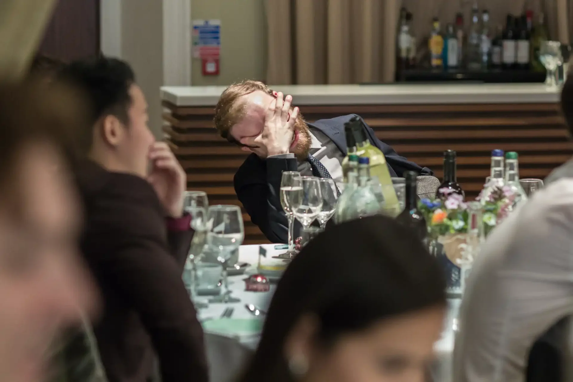 A man with a beard resting his head on his hand appears to be asleep or fatigued at a busy dinner event.