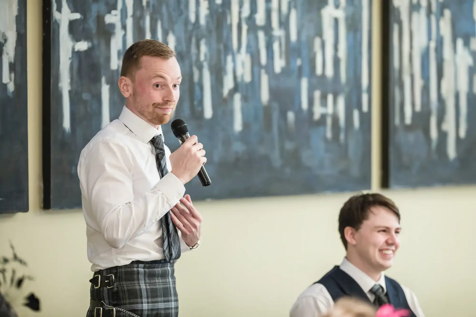 A man in a vest and kilt speaks into a microphone at an event, with a seated man smiling in the background.