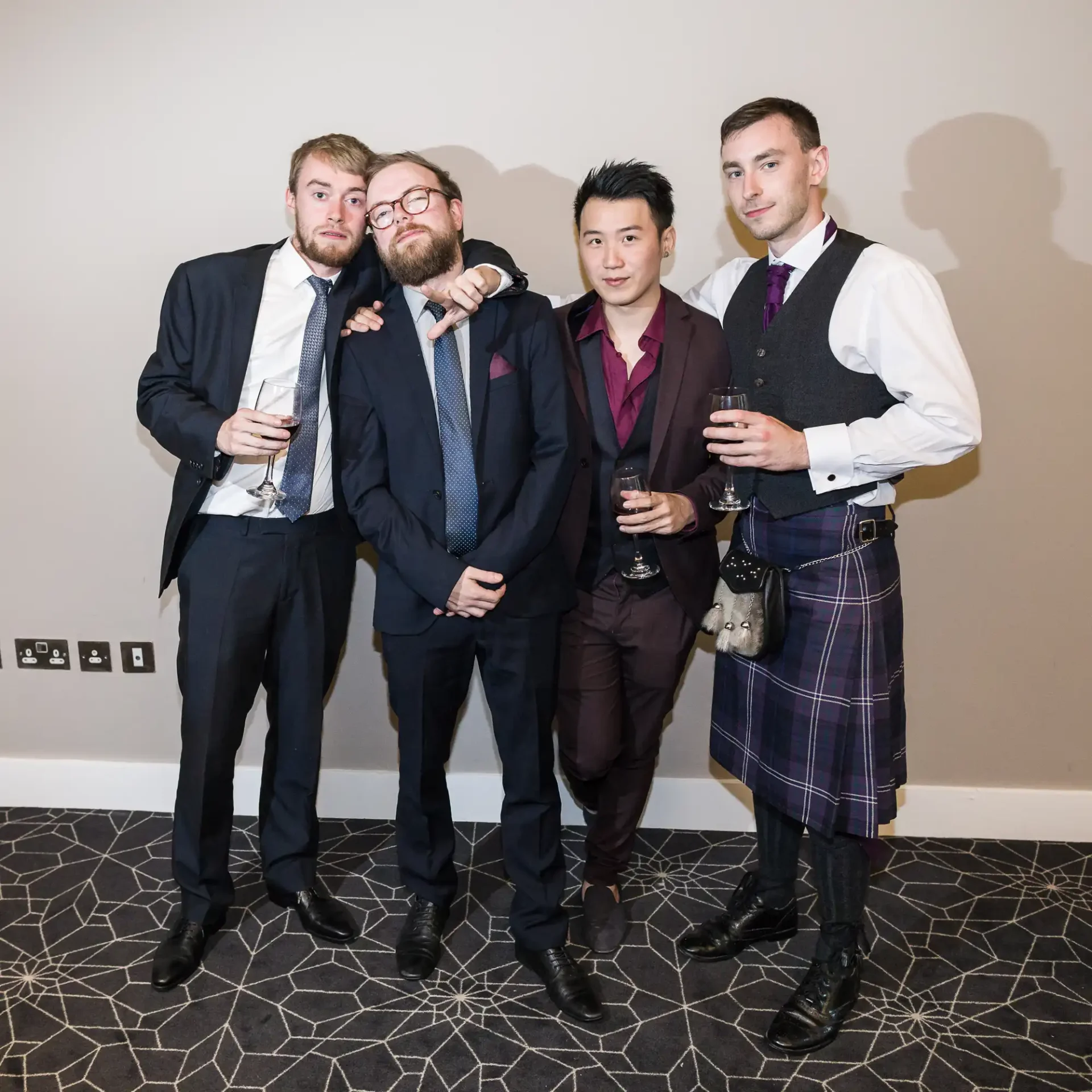 Four men in formal wear, one in a kilt, pose with drinks at an indoor event.