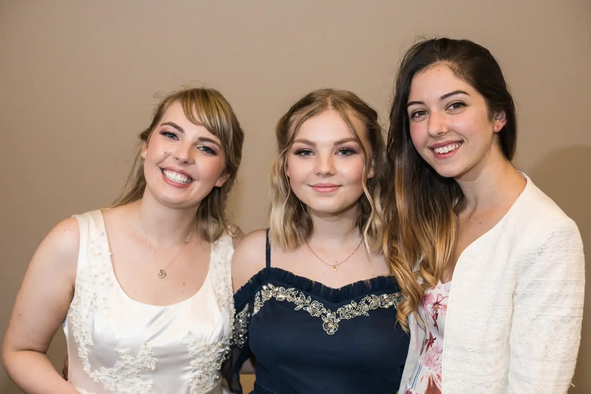 Three young women smiling at the camera, two in white dresses and one in a blue and black dress.