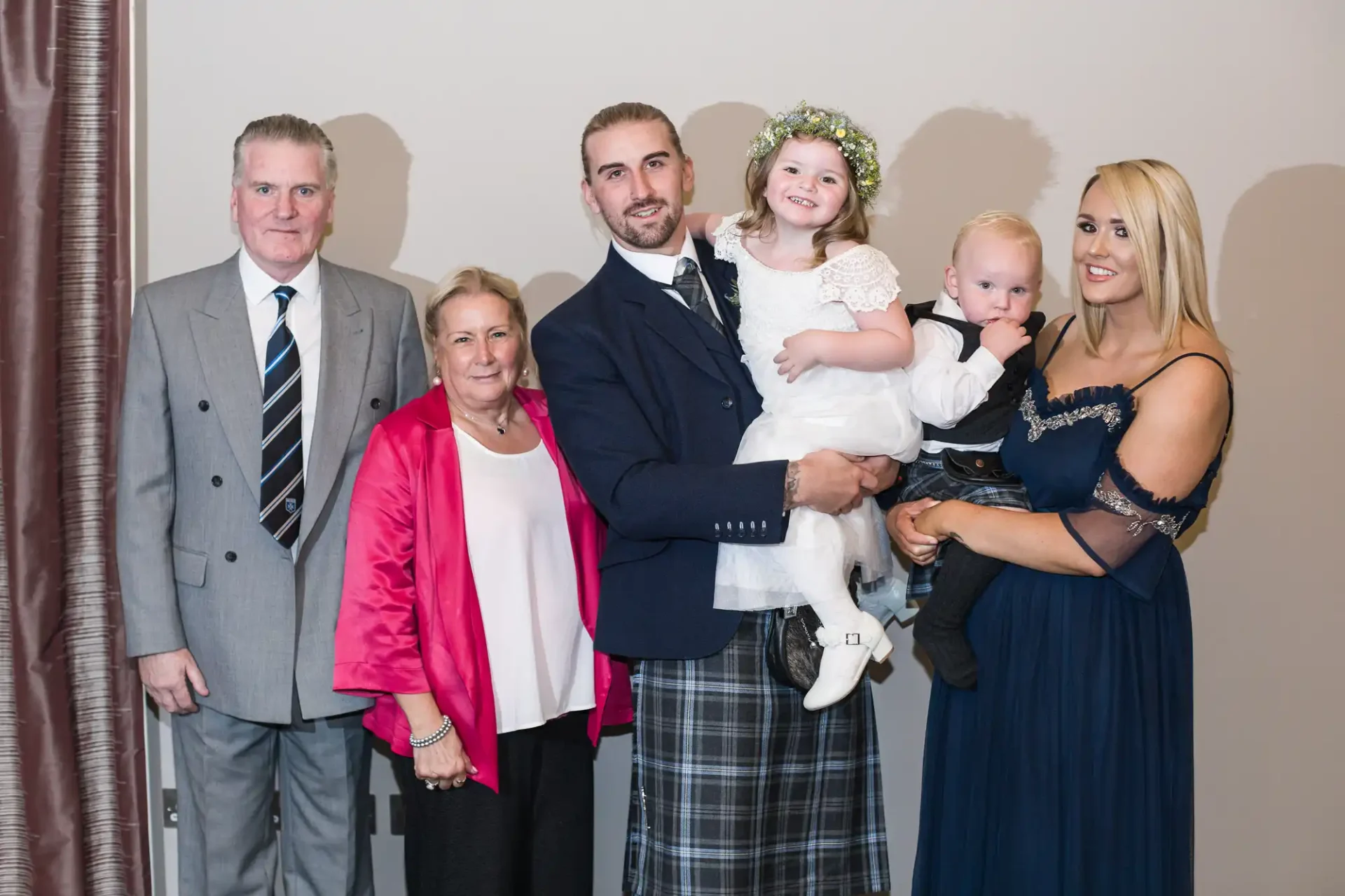 A family with three generations posing together: an elderly couple, a young man in a tartan kilt holding a toddler, a young woman, and a small girl in a floral crown.