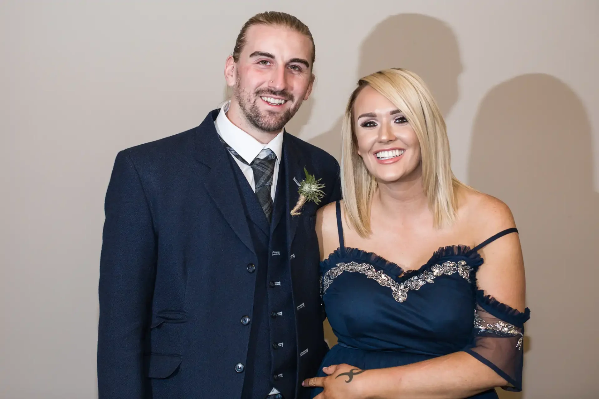 A man in a dark suit with a boutonniere and a woman in a navy blue dress with sequin details, smiling and standing together against a beige wall.