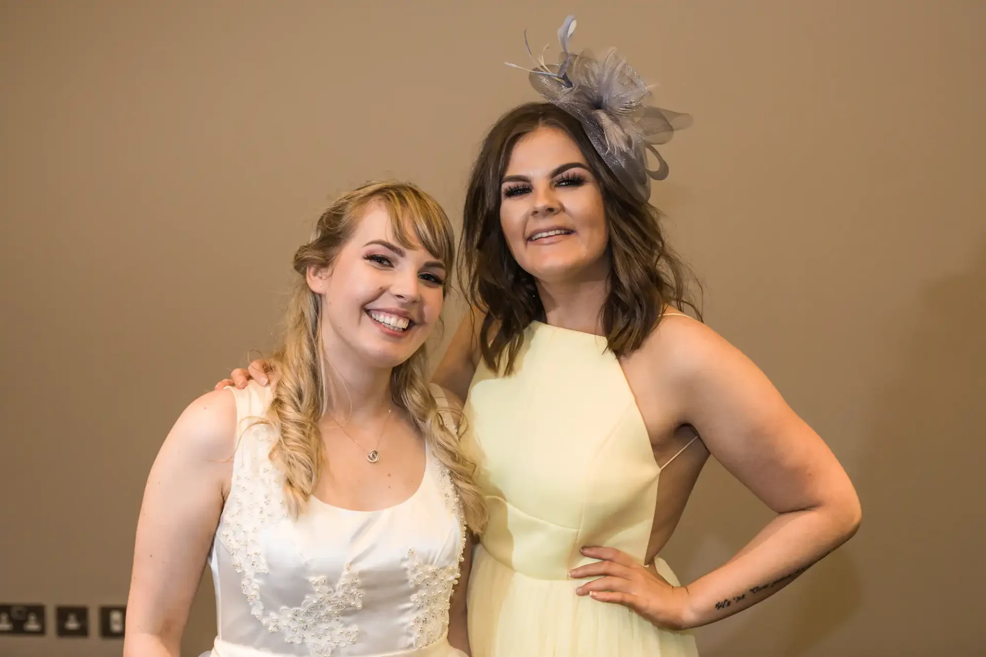 Two women smiling at a formal event, one in a white bridal dress and the other in a yellow dress with a gray fascinator.