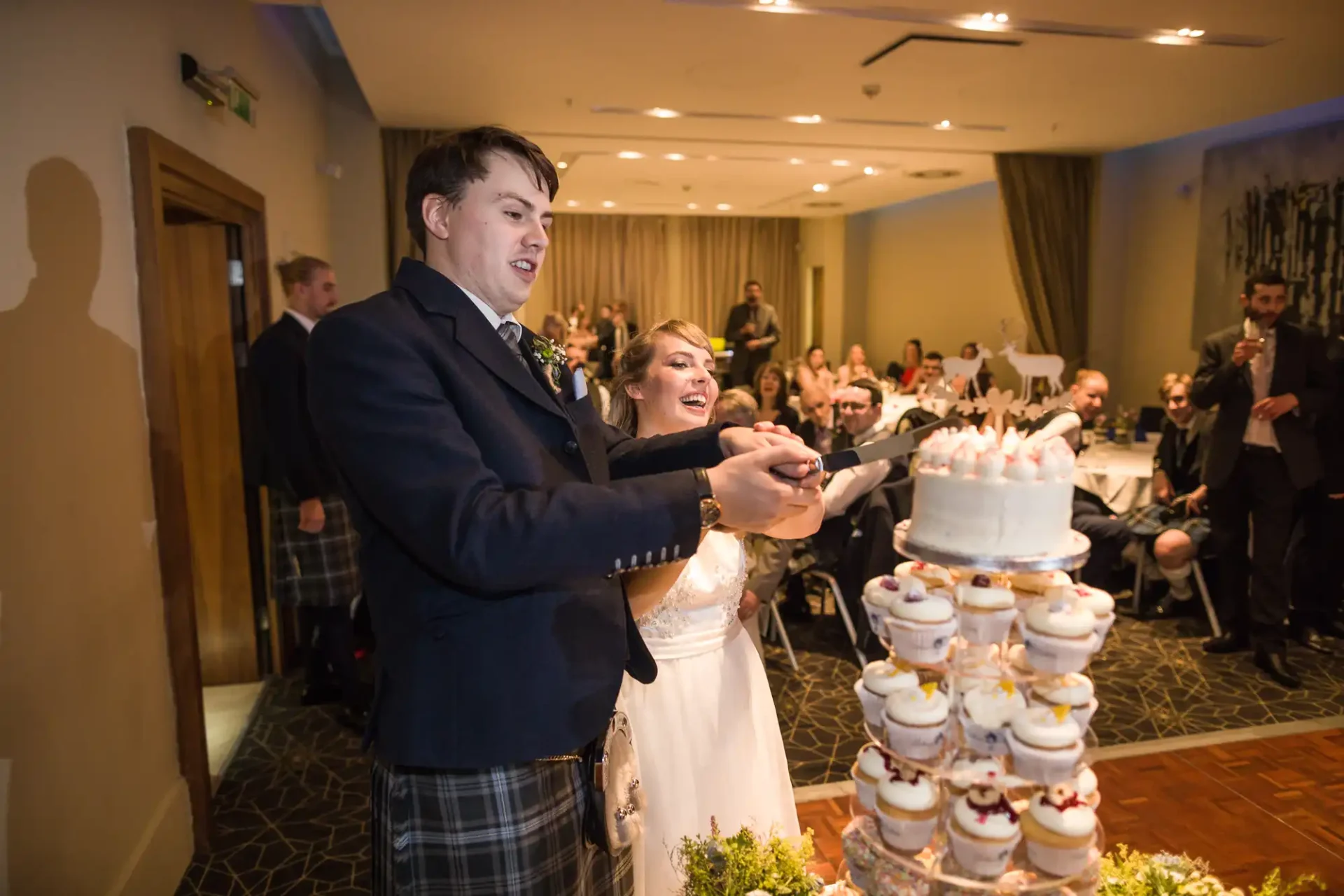Bride and groom in formal attire cutting a tiered wedding cake at a reception hall, surrounded by guests.