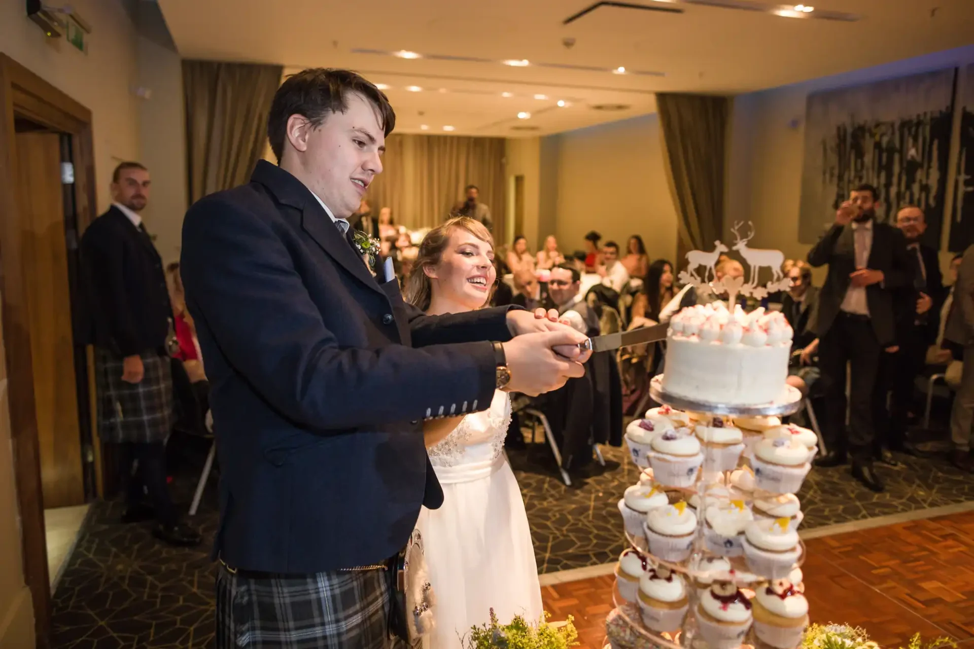 A bride and groom cutting a tiered wedding cake surrounded by guests in a warmly lit room.