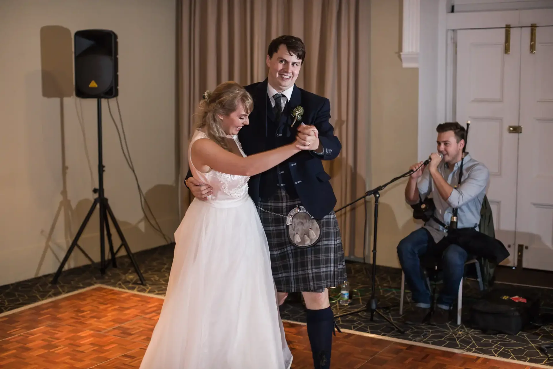 Bride and groom sharing a dance, the groom wearing a kilt, with a musician playing the accordion in the background.