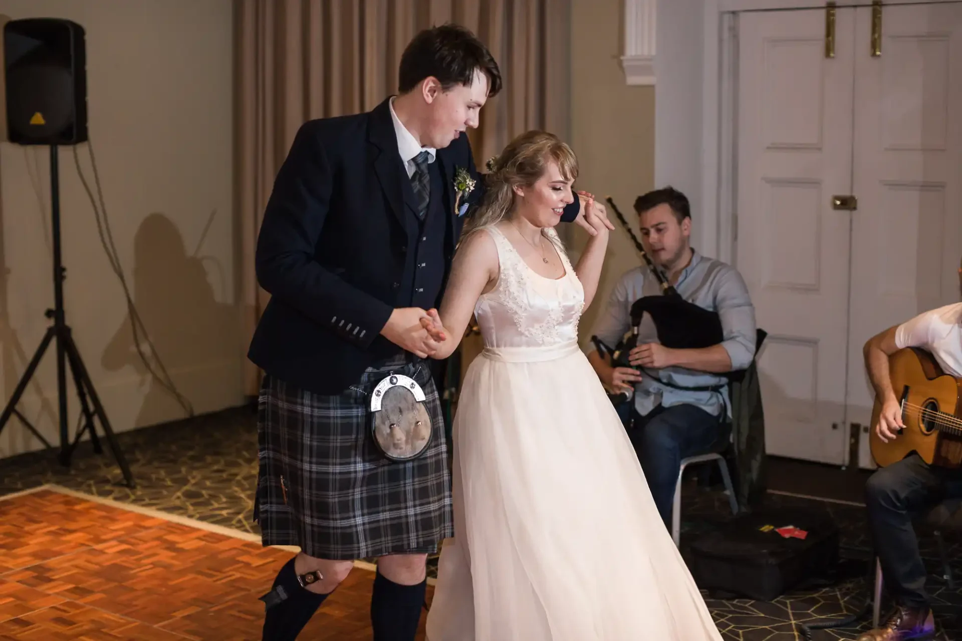 A bride and groom in wedding attire dancing joyfully, the groom wearing a kilt, with musicians playing in the background.