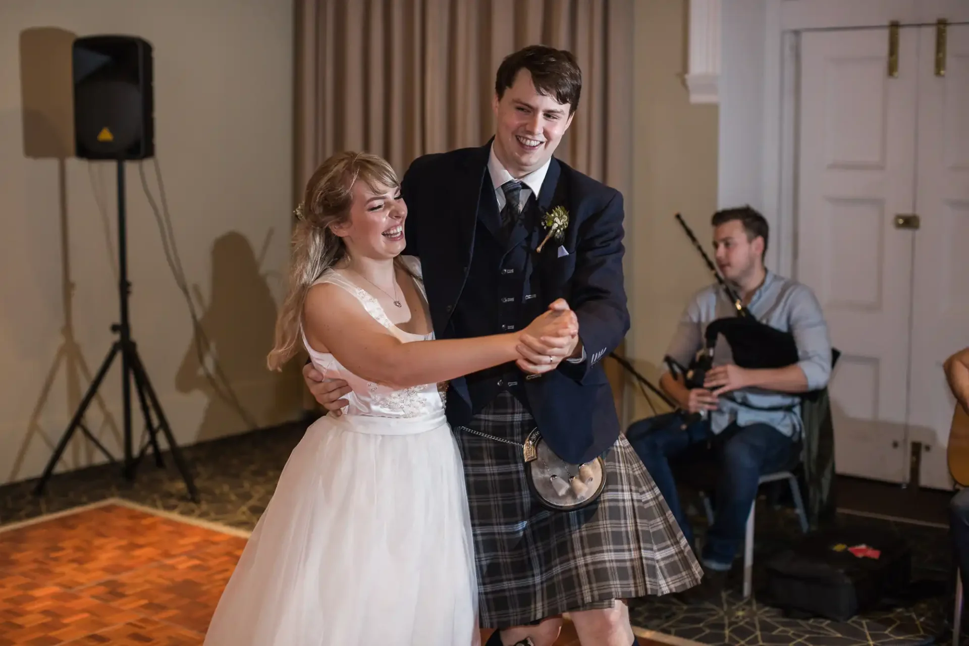 A joyful bride and groom holding hands and dancing at their wedding reception, with a live band playing in the background. the groom is wearing a kilt.