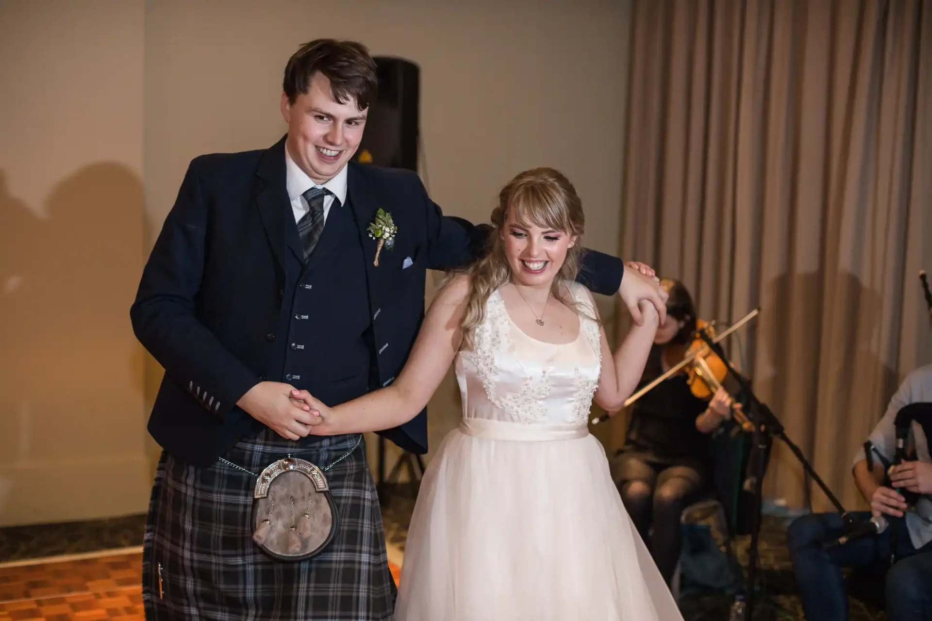 A bride and groom smiling and dancing, the groom wearing a kilt and the bride in a white dress, with a violinist playing in the background.